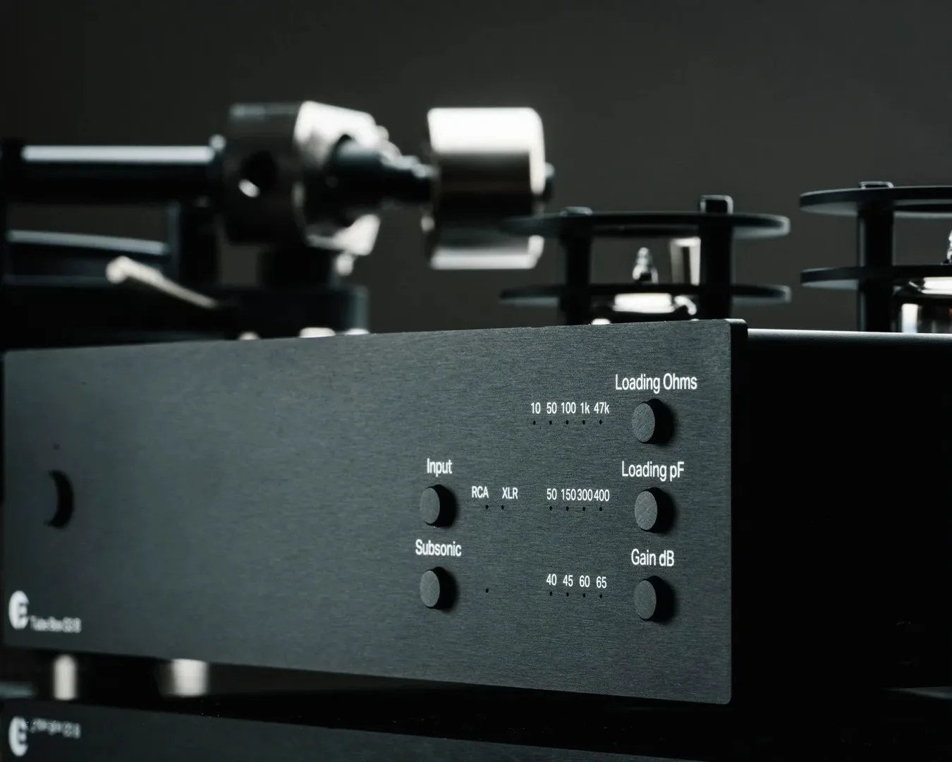 Close-up of a black audio device with multiple control knobs labeled for Input (RCA, XLR), Subsonic, Loading Ohms (10, 50, 100, 1k, 47k), Loading pF (50, 150, 300, 400), and Gain dB (40, 45, 60, 65). The background is dark, and parts of the device's mechanical components are visible but out of focus.