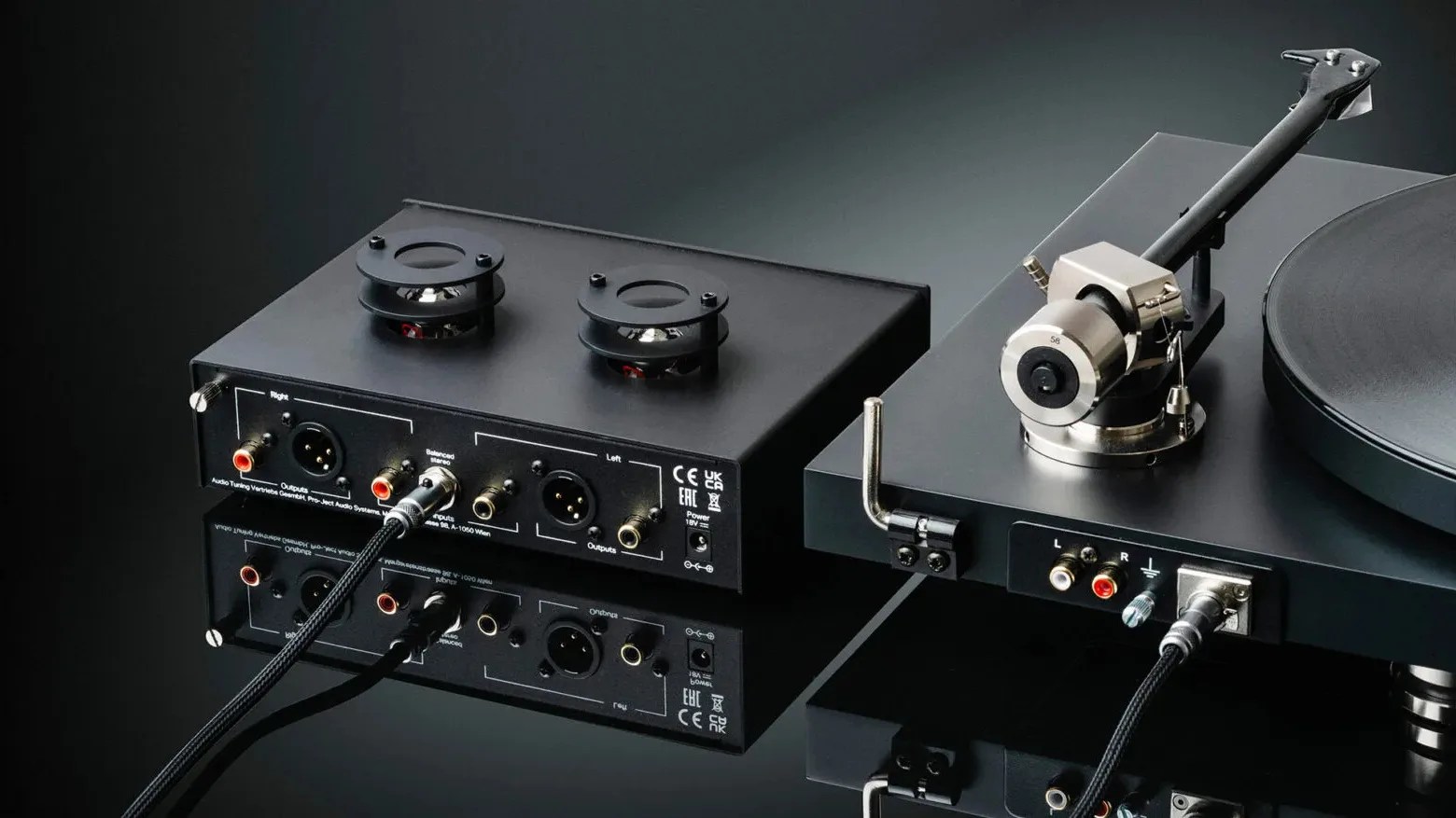 A close-up of a high-end audio setup featuring a black turntable with a precision tonearm and a separate black audio component with two small speakers on top. The audio component has multiple input and output ports, including RCA and XLR connectors, and is connected to the turntable with cables. The setup is placed on a reflective black surface against a dark background.