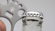 A close-up of a silver bottle opener with an octopus logo being used to pry off a silver bottle cap from a glass bottle.