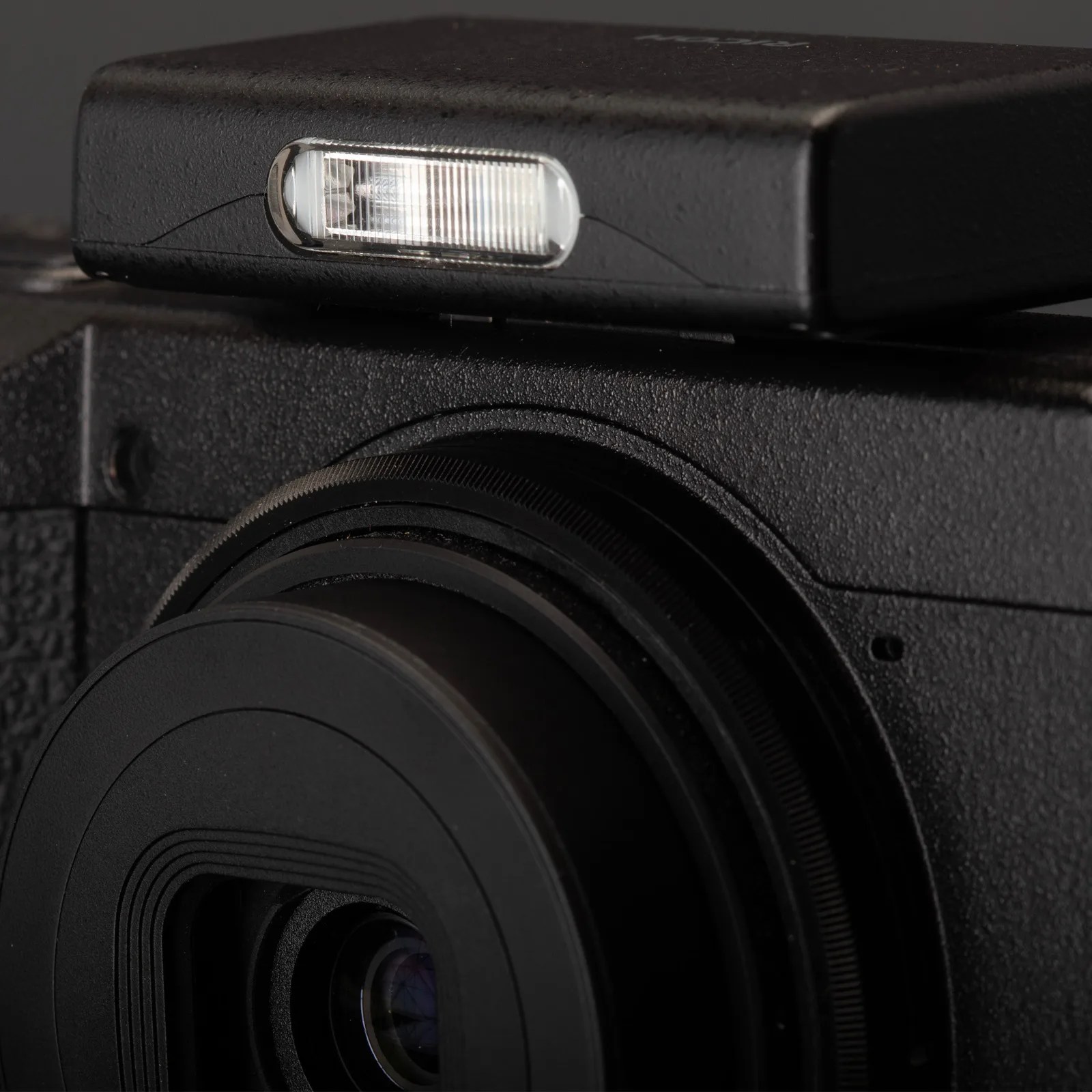 Close-up of a black camera lens with a lens cap on, showing the textured body of the camera and a rectangular flash unit positioned on top. The camera and flash have a matte black finish.
