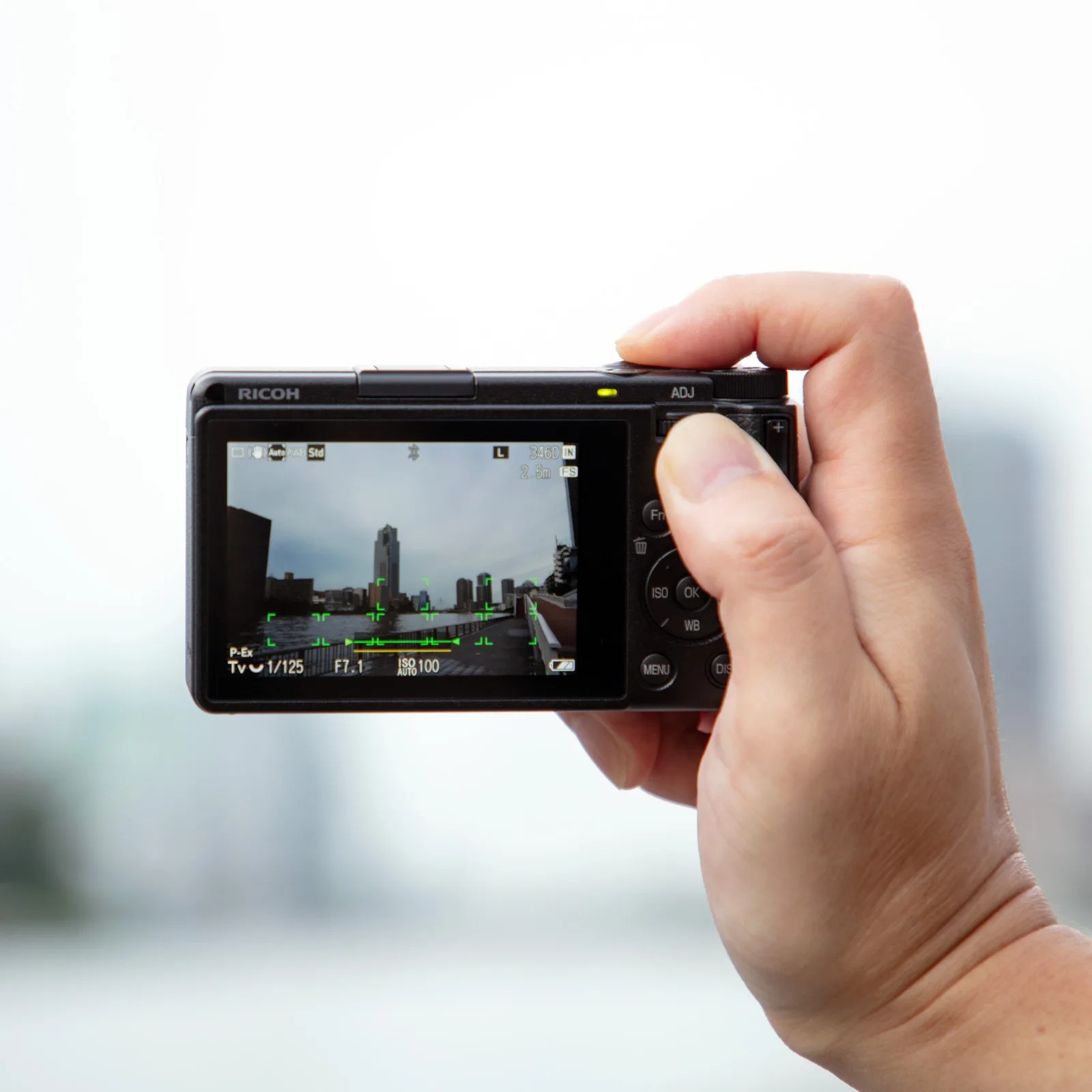 Hand holding a Ricoh digital camera, showing the camera's screen displaying a cityscape with buildings and a river under a cloudy sky. The camera settings visible include shutter speed 1/125, aperture f/7.1, and ISO 100.