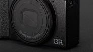 Close-up of a black camera with a textured body, featuring a GR lens with specifications "f=18.3mm 1:2.8" and the letters "GR" prominently displayed on the lower right corner of the camera body.