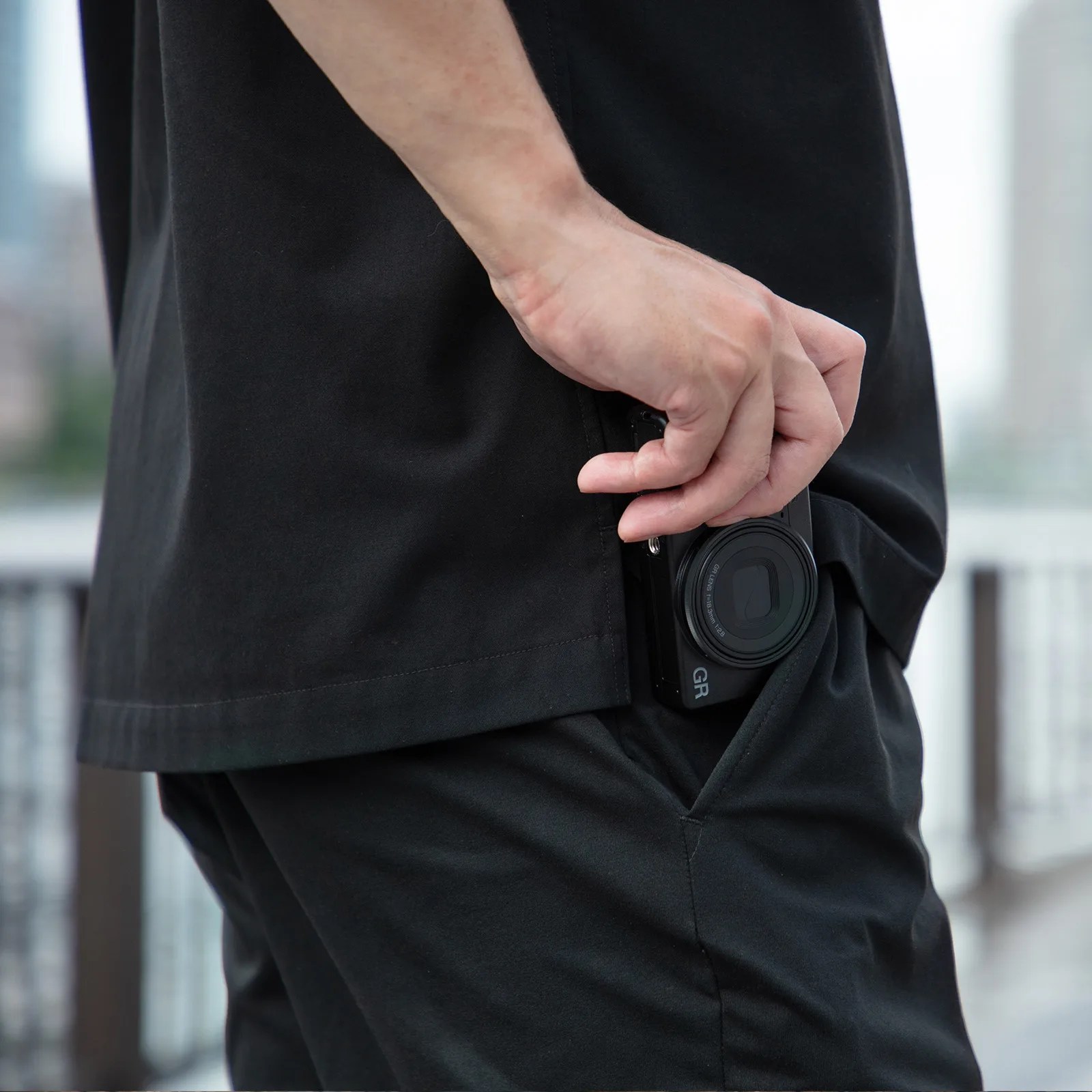 A person wearing black clothing is holding a compact black camera labeled "GR" partially inside their right pocket. The background is blurred, showing an outdoor setting with railings.