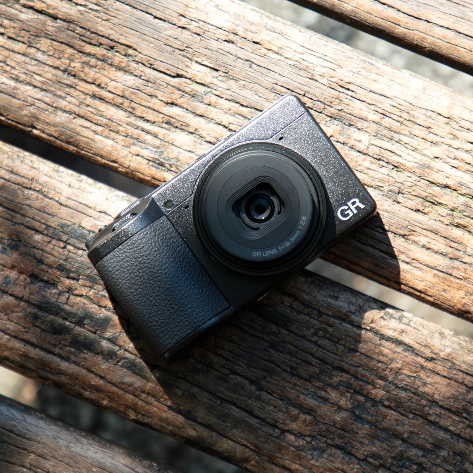 Compact black digital camera with a textured grip, labeled "GR" on the front, resting on a weathered wooden surface with sunlight casting shadows. The lens is centered and marked with "GR LENS f=18.3mm 1:2.8".