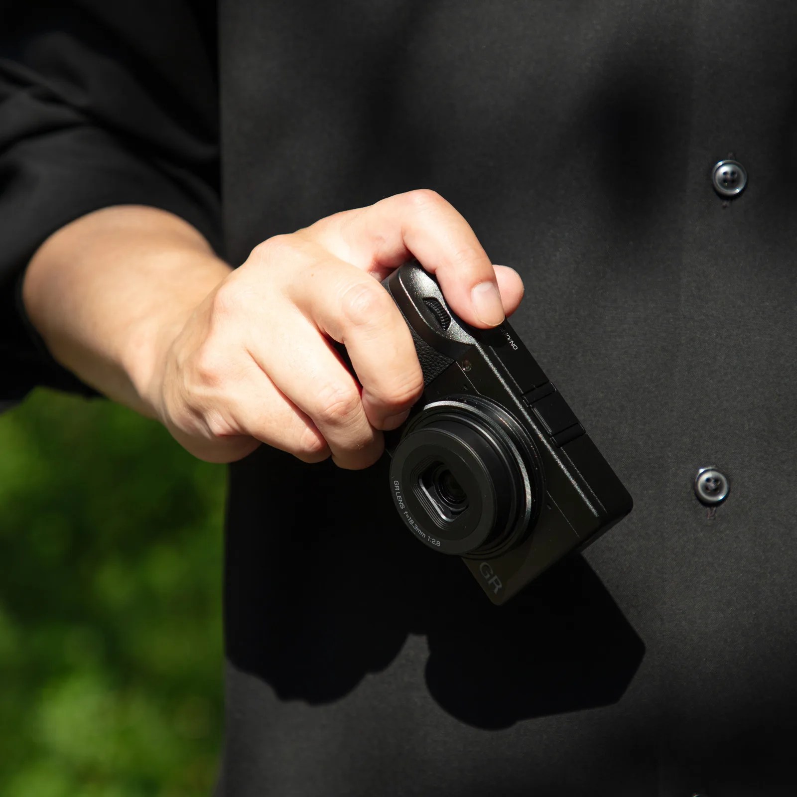 A person wearing a black shirt is holding a compact black digital camera with a visible lens marked "GR LENS 18.3mm 1:2.8." The camera is held in the right hand against a blurred green background.