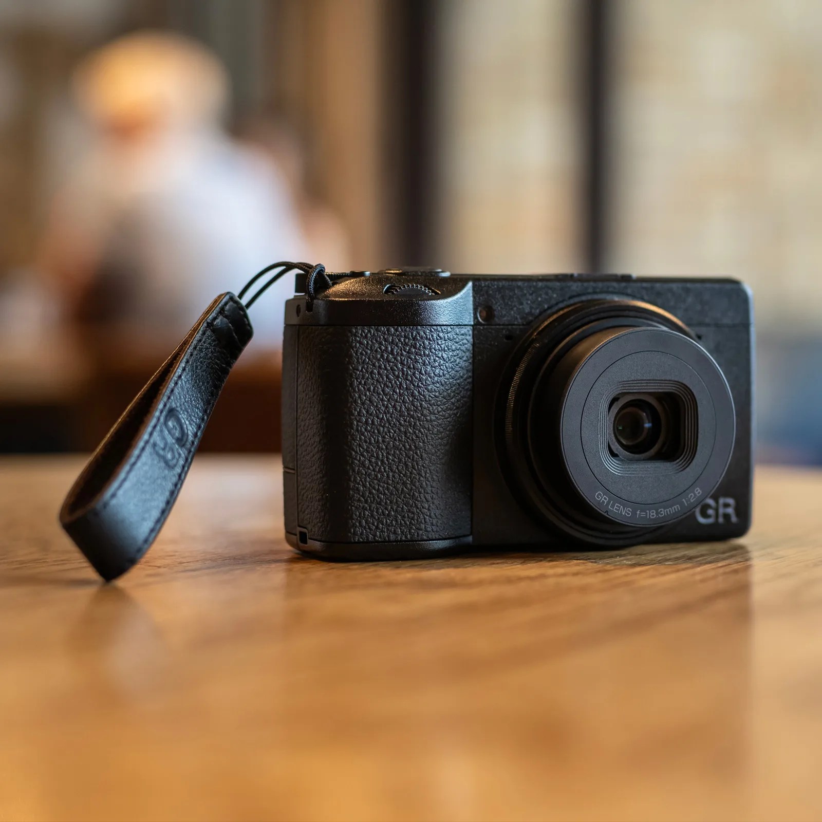 Compact black digital camera with a textured grip, a prominent lens marked "GR LENS f=18.3mm 1:2.8," and a black wrist strap attached, placed on a wooden surface with a blurred background.
