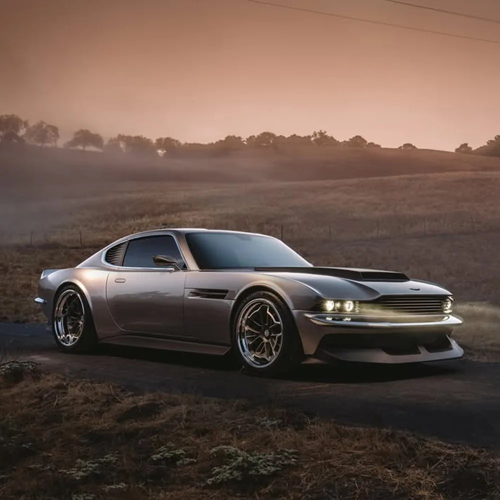 Sleek silver sports car with a low profile and wide body, featuring modern LED headlights and a prominent front grille, parked on a rural road at dusk with a misty, tree-lined hillside in the background.