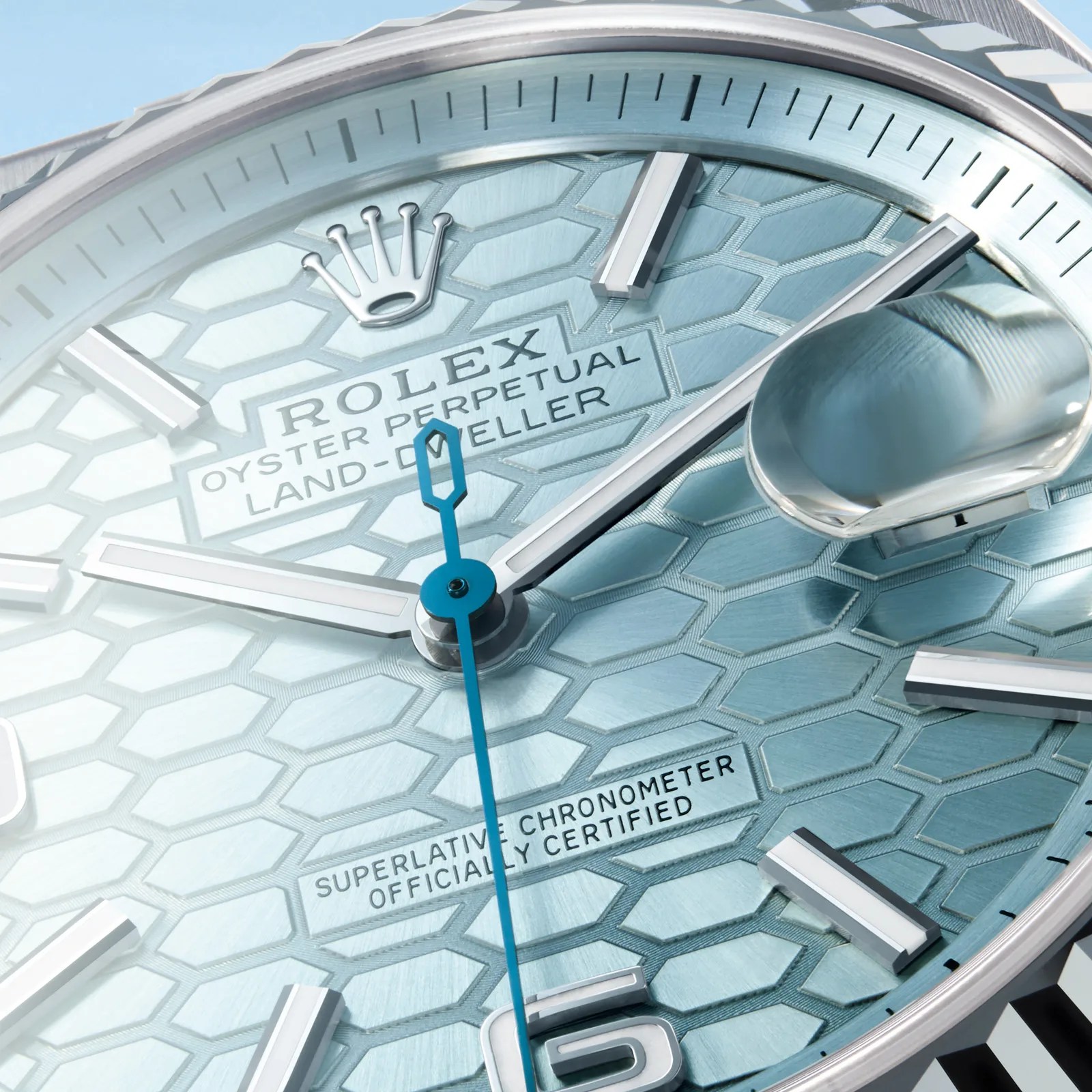 Close-up of a Rolex Oyster Perpetual Land-Dweller watch face with a light blue textured dial, silver hour markers, and hands. The second hand is blue, and there is a magnified date window on the right side. The text "ROLEX OYSTER PERPETUAL LAND-DWELLER" and "SUPERLATIVE CHRONOMETER OFFICIALLY CERTIFIED" is visible on the dial.