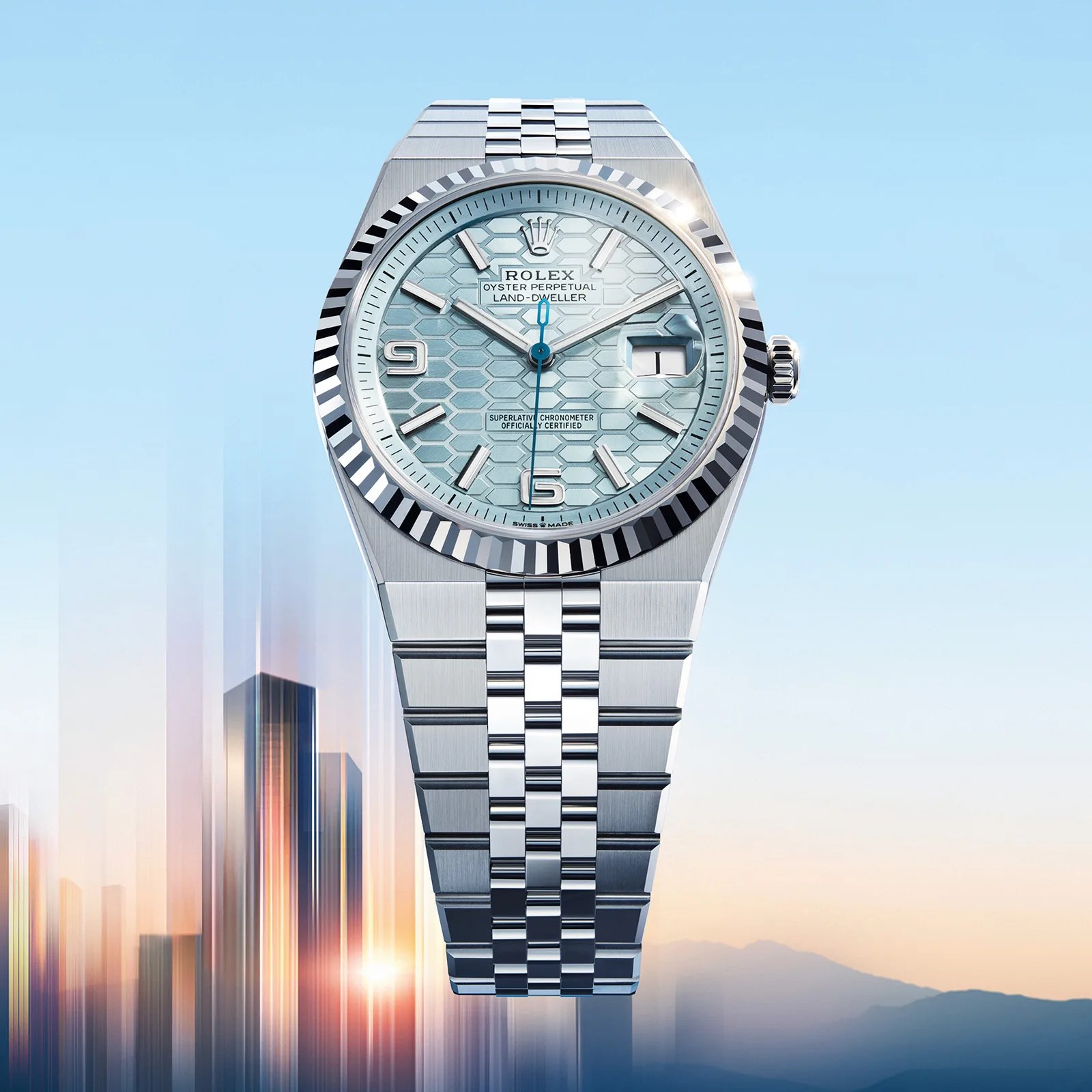 Rolex Oyster Perpetual Land-Dweller watch with a silver metal bracelet and a light blue hexagonal patterned dial. The watch features silver hour markers, a date window at 3 o'clock with a magnifying lens, and a fluted bezel. The background shows a blurred cityscape with a sunrise or sunset sky.