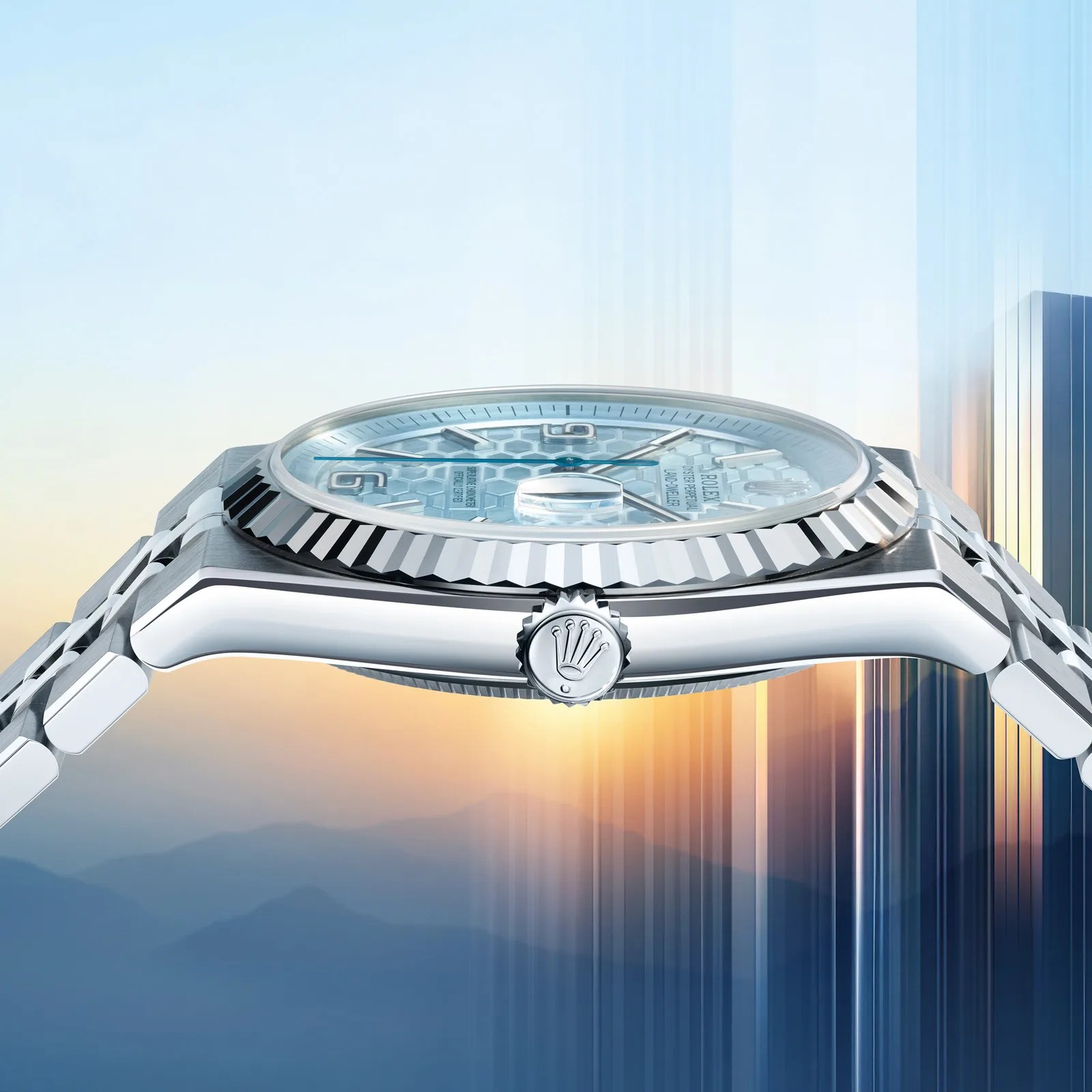 Side view of a silver Rolex watch with a fluted bezel and a light blue honeycomb-patterned dial. The watch features a polished metal bracelet and a crown with the Rolex logo. The background shows a blurred gradient of blue and orange tones resembling a sunrise or sunset over mountains.