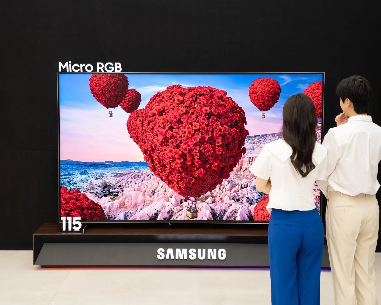 A large Samsung Micro RGB 115-inch television displaying a vibrant image of red rose-shaped hot air balloons floating over a rocky landscape. Two people, a woman in a white top and blue pants and a man in a white shirt and beige pants, are standing in front of the TV, viewing the display. The TV is placed on a modern black and wooden stand with the Samsung logo illuminated on the front.