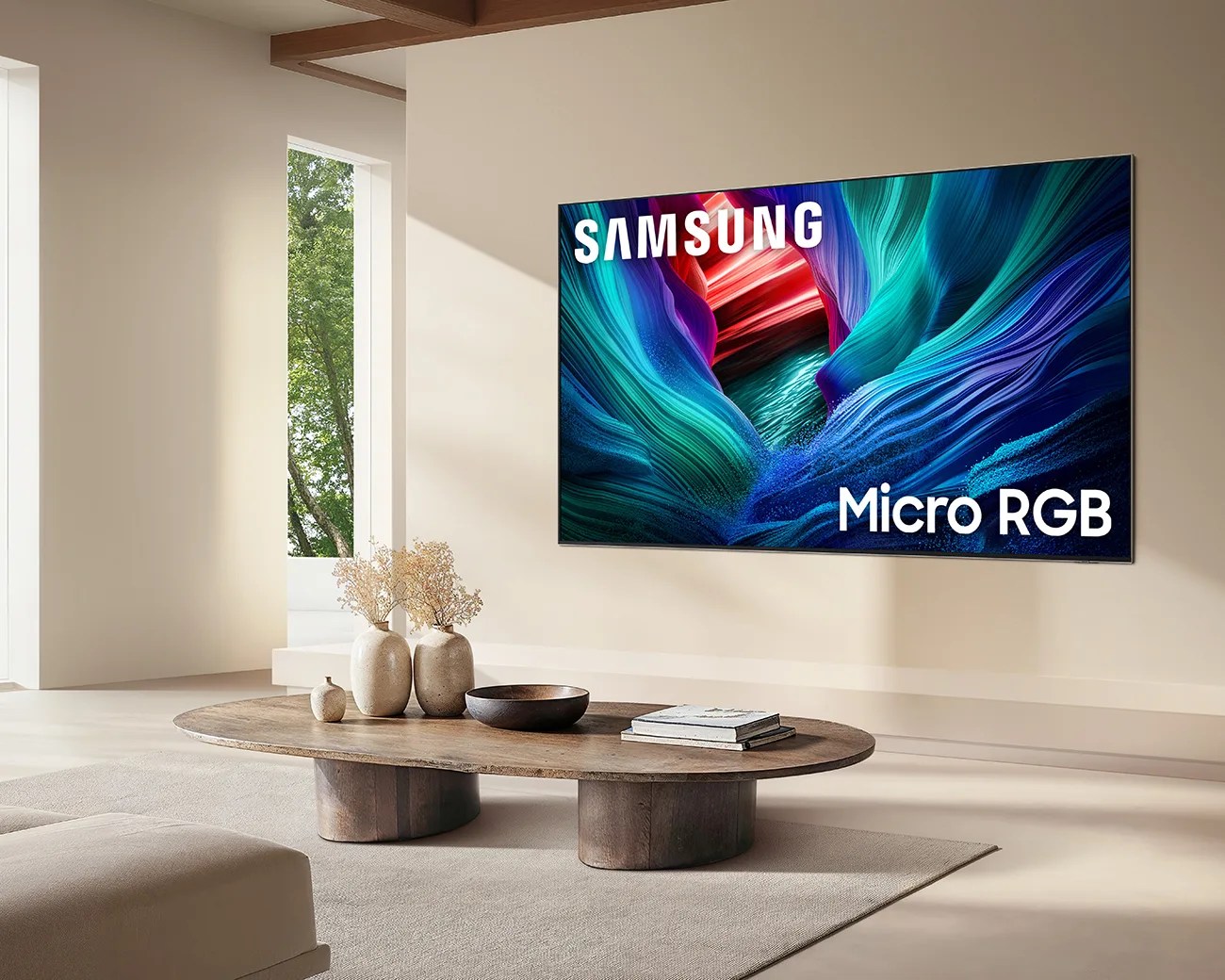 A modern living room with a large Samsung Micro RGB TV mounted on a beige wall. Below the TV is a wooden oval coffee table with two beige vases holding dried plants, a small decorative object, a dark bowl, and two stacked books. The room features neutral tones and natural light coming through a tall window with a view of green trees.