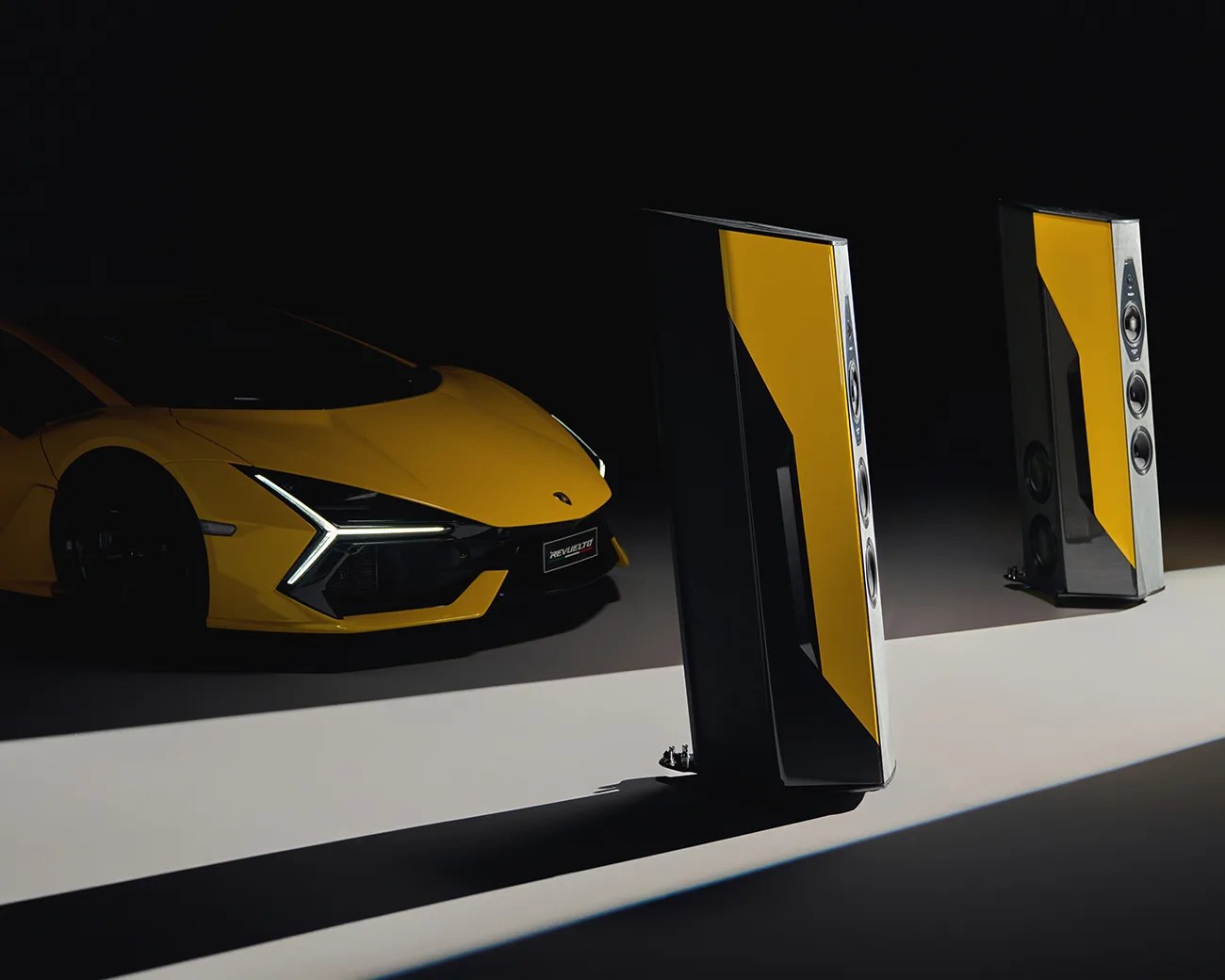 Two tall, modern speakers with yellow and black panels are positioned on a light surface, casting long shadows. In the background, a yellow sports car with distinctive angular headlights is partially visible in the dark.