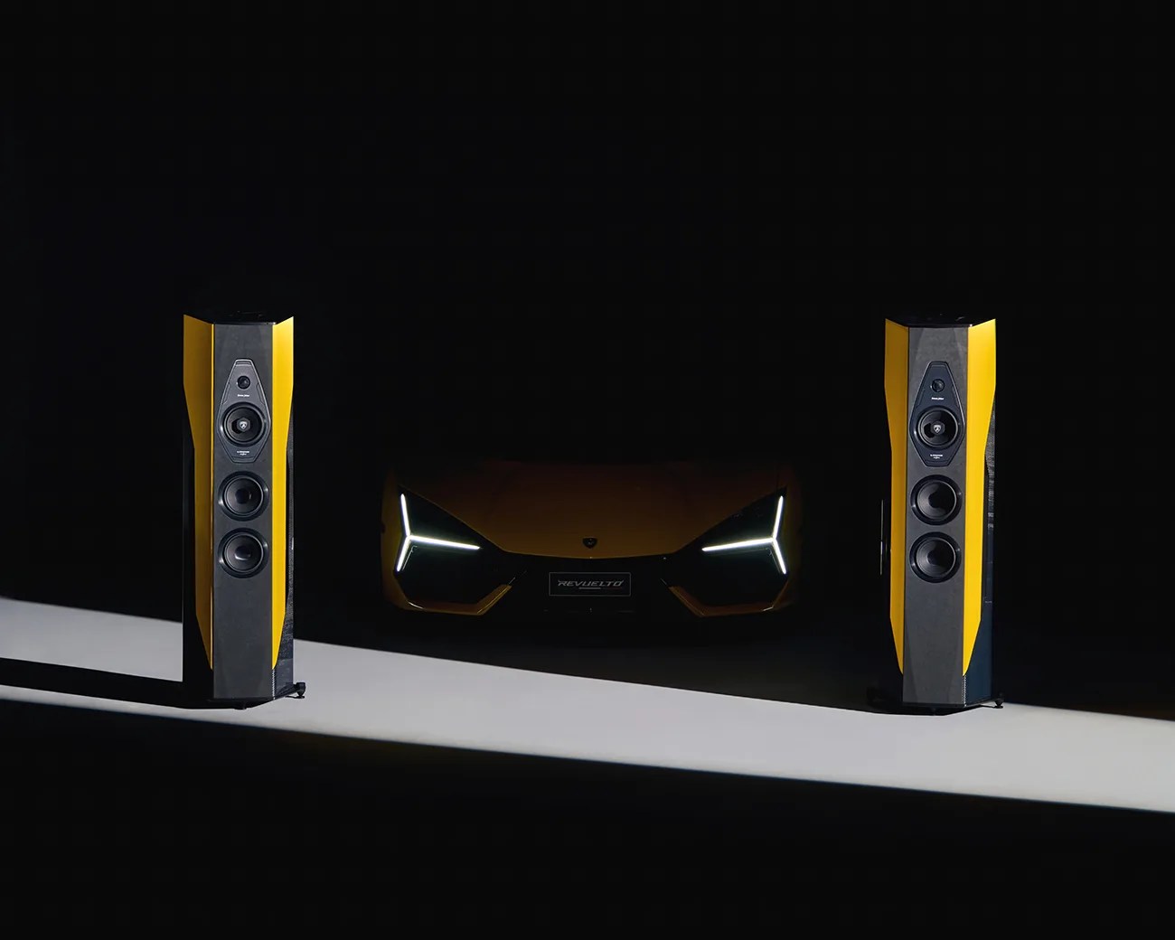 Two tall, yellow and black speakers are positioned on either side of a yellow sports car with distinctive, angular LED headlights glowing in the dark. The car's front grille and license plate are partially visible in the shadowy background.