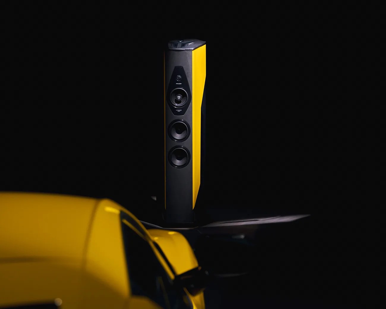 Tall, modern speaker with a black front and yellow sides, positioned against a dark background. The speaker features multiple drivers, including a tweeter, midrange, and two woofers. A yellow car is partially visible in the foreground, matching the speaker's yellow color.