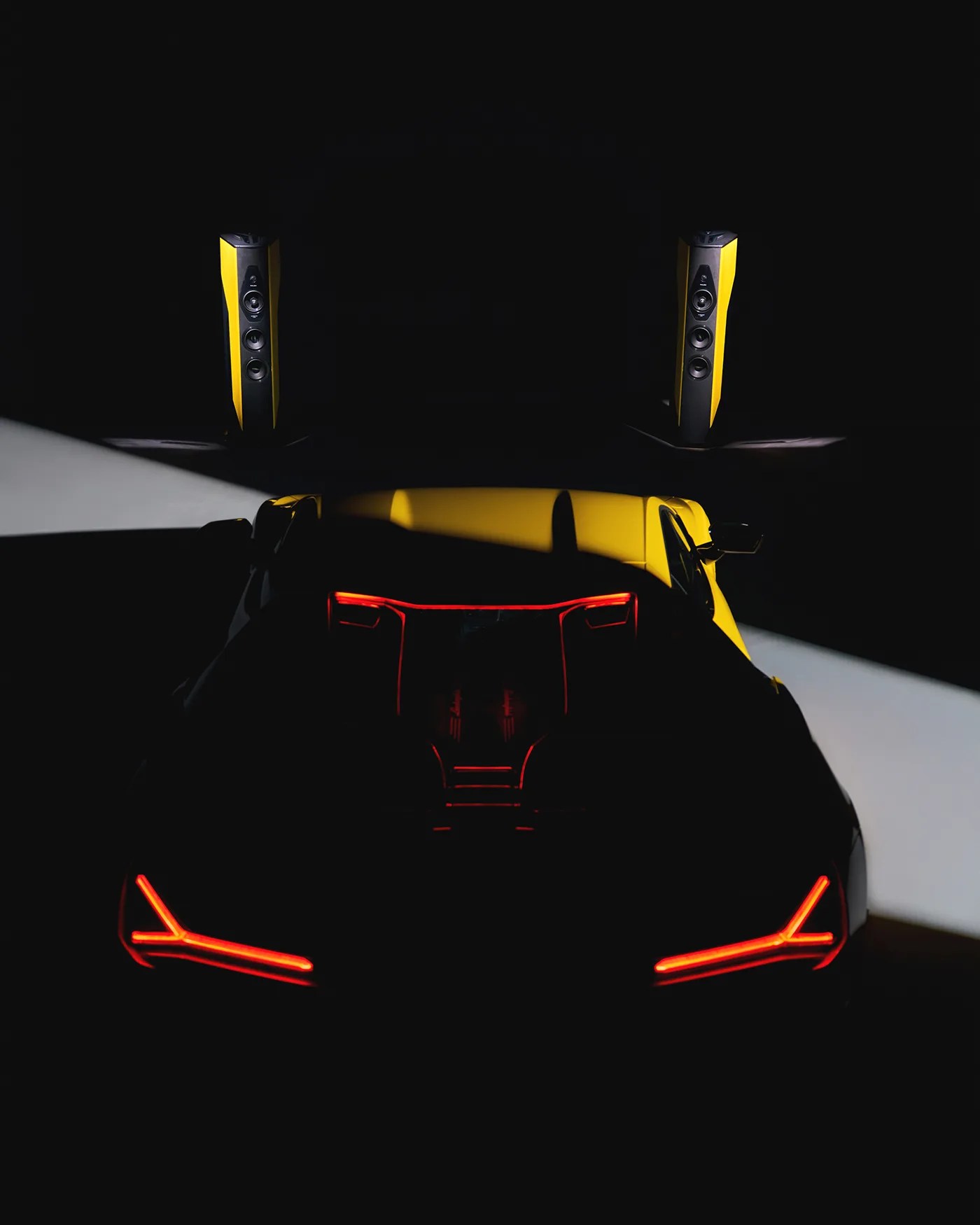 A yellow sports car viewed from the rear in low light, with distinctive red LED taillights illuminated. Two tall, yellow and black speakers are positioned in the background, also highlighted by focused lighting. The scene is predominantly dark, emphasizing the car's sleek lines and the speakers' design.