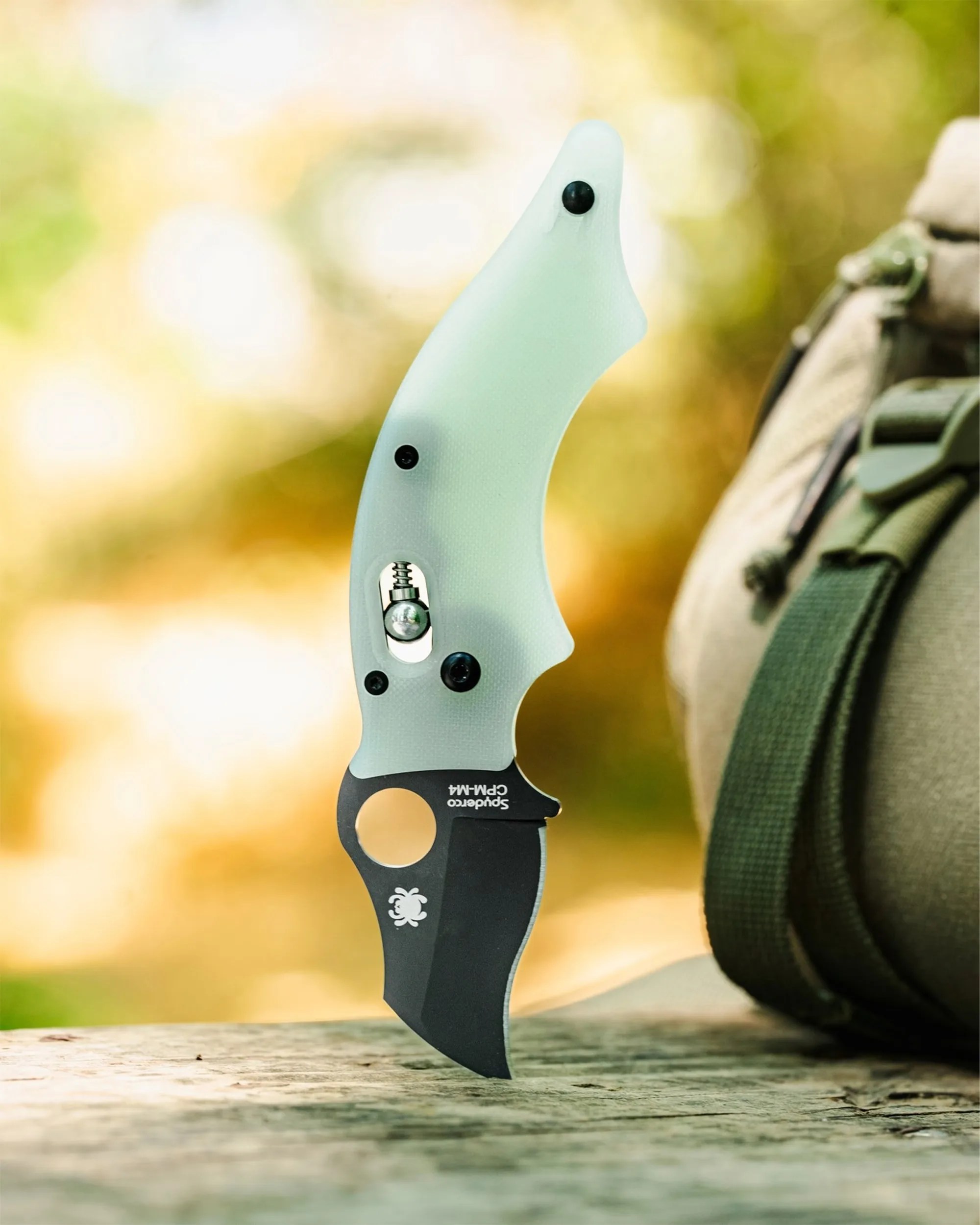 A folding knife with a curved, translucent light green handle and a black blade. The blade has a hole near the base and features the Spyderco logo and "Spyderco CPM-M4" text. The knife is standing upright on a wooden surface with a blurred outdoor background and part of a green and beige backpack visible on the right side.