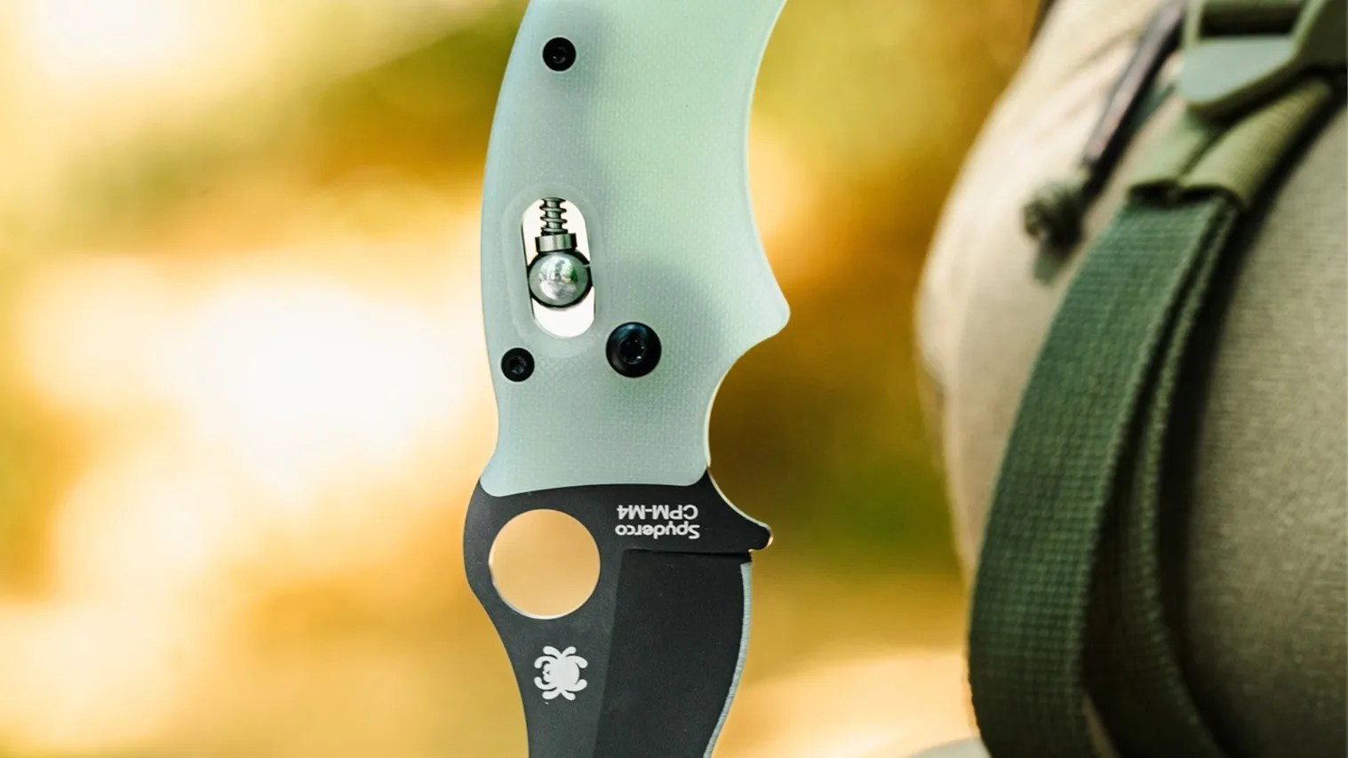 Close-up of a Spyderco folding knife with a light green textured handle and a black blade. The blade features the Spyderco logo and the text "Spyderco CPM-M4." The knife is held next to a green strap and beige fabric background, with a blurred warm-toned background.