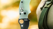 Close-up of a Spyderco folding knife with a light green textured handle and a black blade. The blade features the Spyderco logo and the text "Spyderco CPM-M4." The knife is held next to a green strap and beige fabric background, with a blurred warm-toned background.