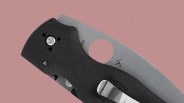 Close-up of a folding knife with a black textured handle and a silver blade. The blade features a round hole near the base and has markings including "Golden" The handle has a metal clip attached. The background is a muted pink color.