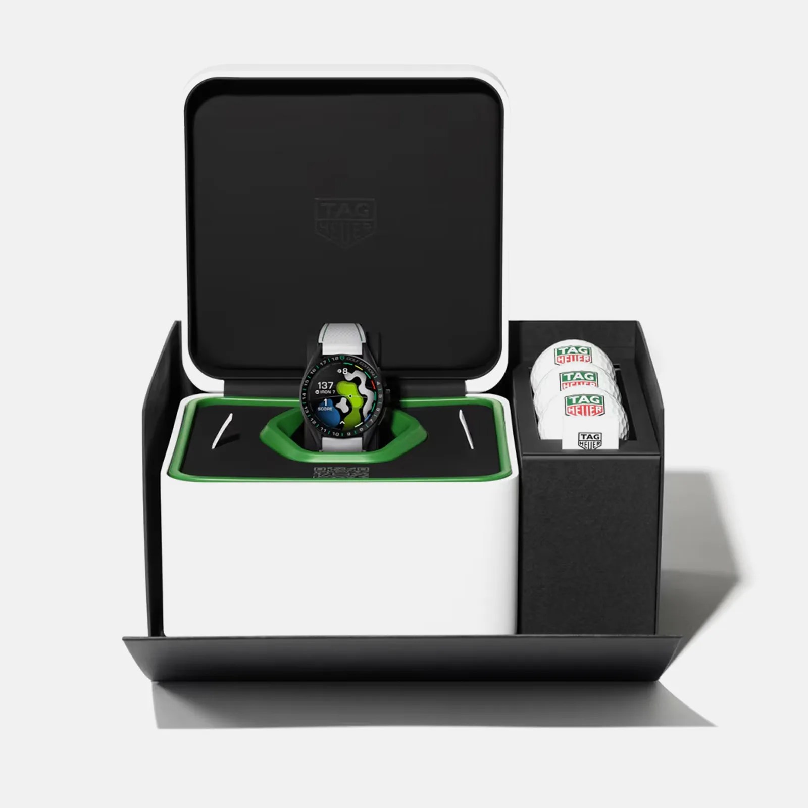 A TAG Heuer smartwatch with a white strap displayed in an open black and white box. The watch face shows a colorful golf course map and various golf-related metrics. Next to the watch, there is a compartment holding three white golf balls with the TAG Heuer logo. The interior of the box has green accents around the watch.