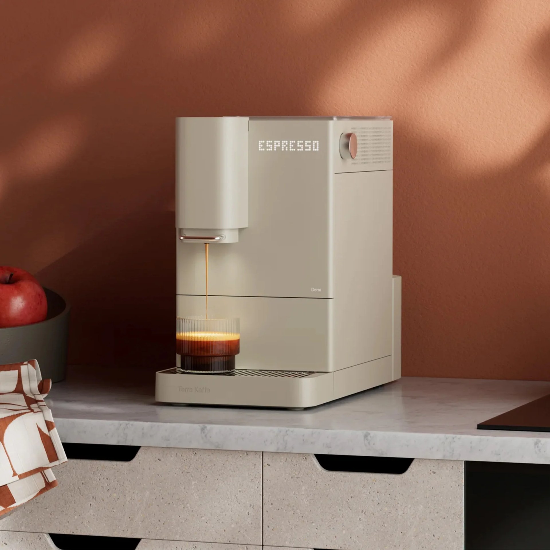 A modern, beige espresso machine labeled "Terra Kaffa" is placed on a marble countertop. The machine is dispensing espresso into a clear, ribbed glass cup. The word "ESPRESSO" is displayed in white digital text on the front of the machine. The background features a warm, brown textured wall, and a bowl with a red apple is partially visible on the left side. A patterned cloth with brown and white colors is draped over the edge of the countertop.