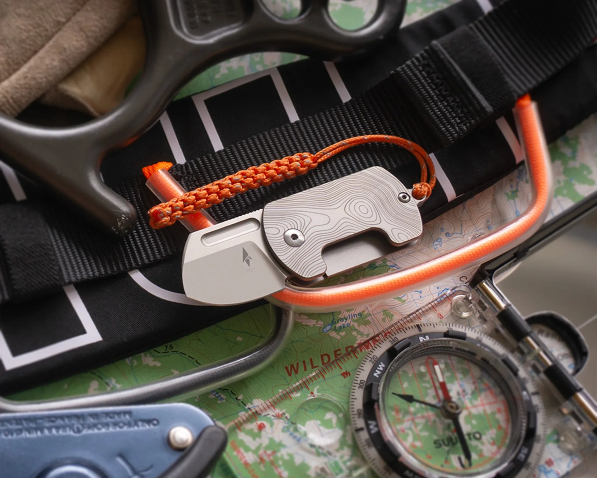 A small folding knife with a silver blade and a handle featuring a topographic map pattern, attached to an orange braided lanyard. The knife is placed on a black strap over a green topographic map, alongside a clear compass with a black and red needle, and other outdoor gear.