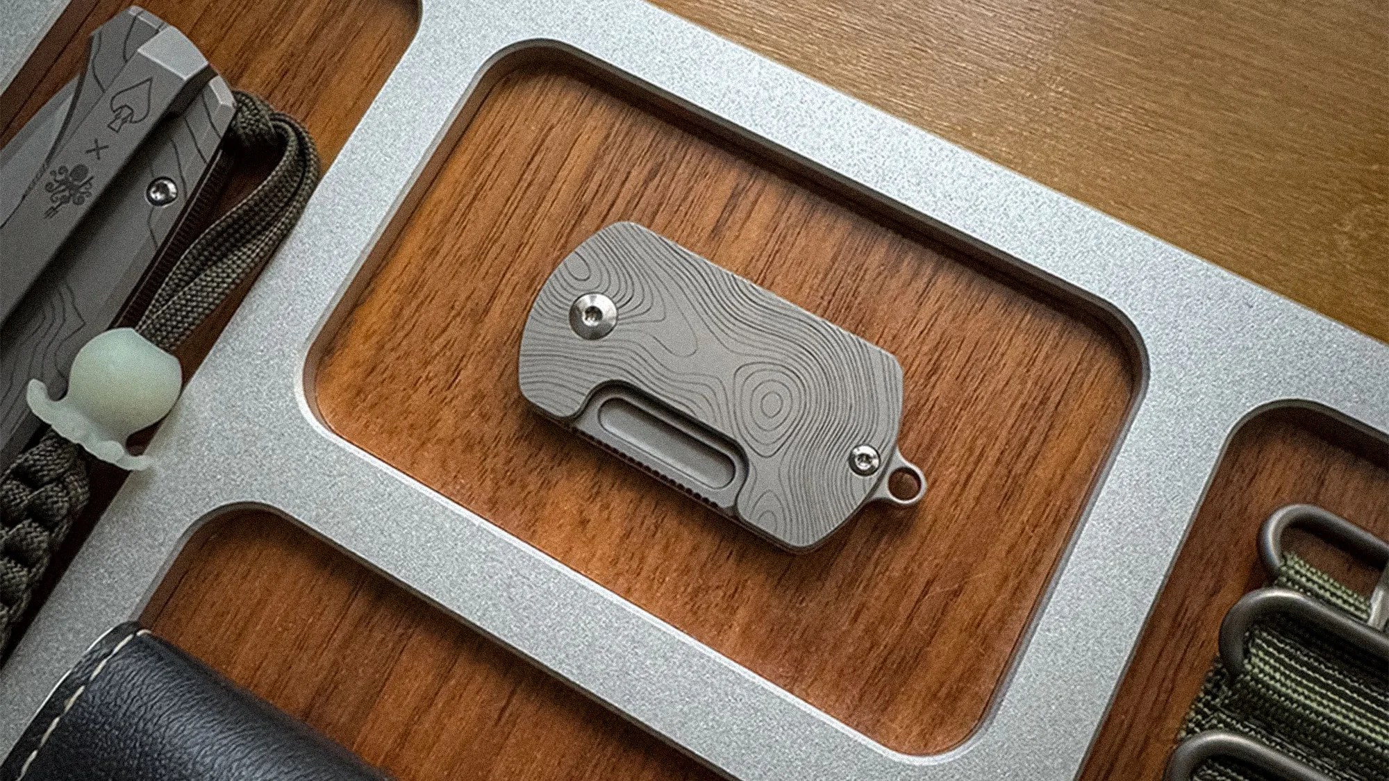 A small, rectangular metal object with a topographic pattern engraved on its surface, secured with two screws, and featuring a small loop on one end. It is placed in a recessed compartment of a silver metal tray with a wooden base.