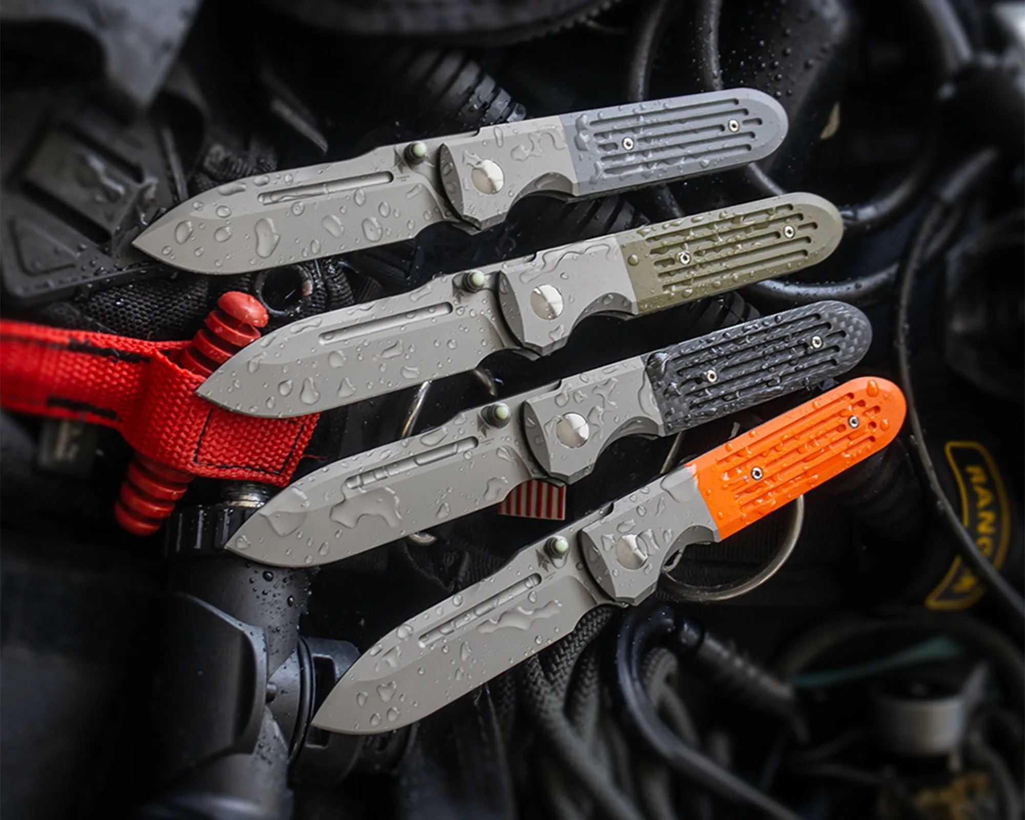 Four folding knives with water droplets on their blades and handles are arranged in a row. Each knife has a gray blade and a handle with a different color section: light gray, olive green, black, and bright orange. The knives are placed on a dark, textured surface with various cables and equipment around them.