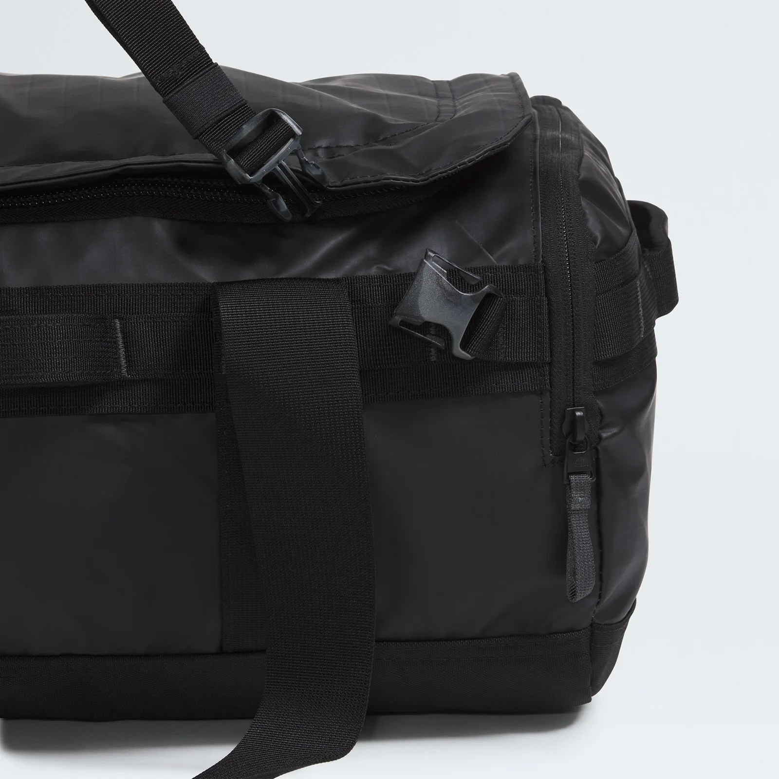 Close-up of a black bag showing a zipper on the right side, a black plastic buckle, and a wide black strap. The bag material appears smooth and slightly shiny, with reinforced stitching and webbing details.