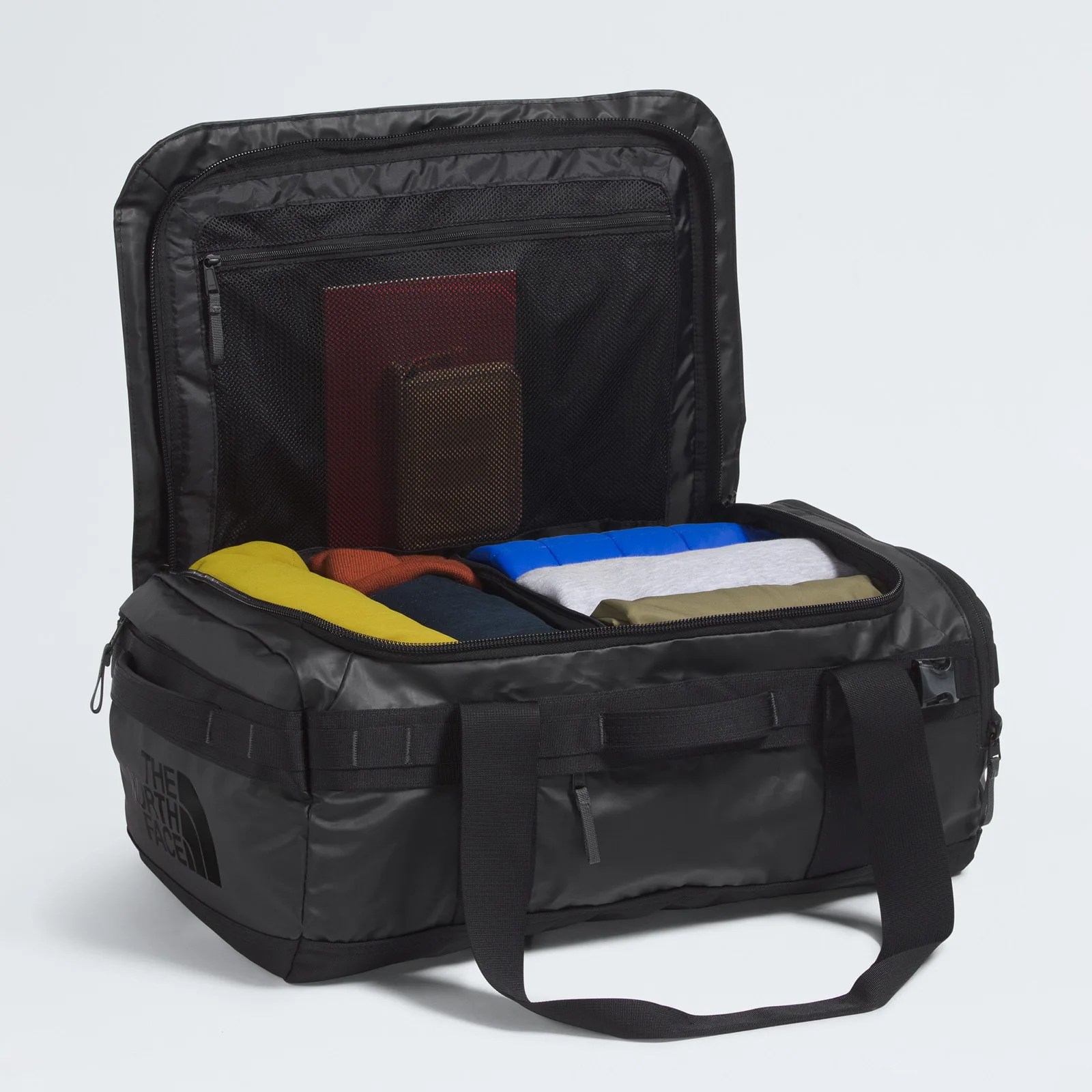 Black rectangular duffel bag with a zippered lid open, showing neatly folded colorful clothes inside. The interior lid has a mesh zippered pocket containing a red notebook and a brown wallet. The bag has black handles and "The North Face" logo printed on the side.