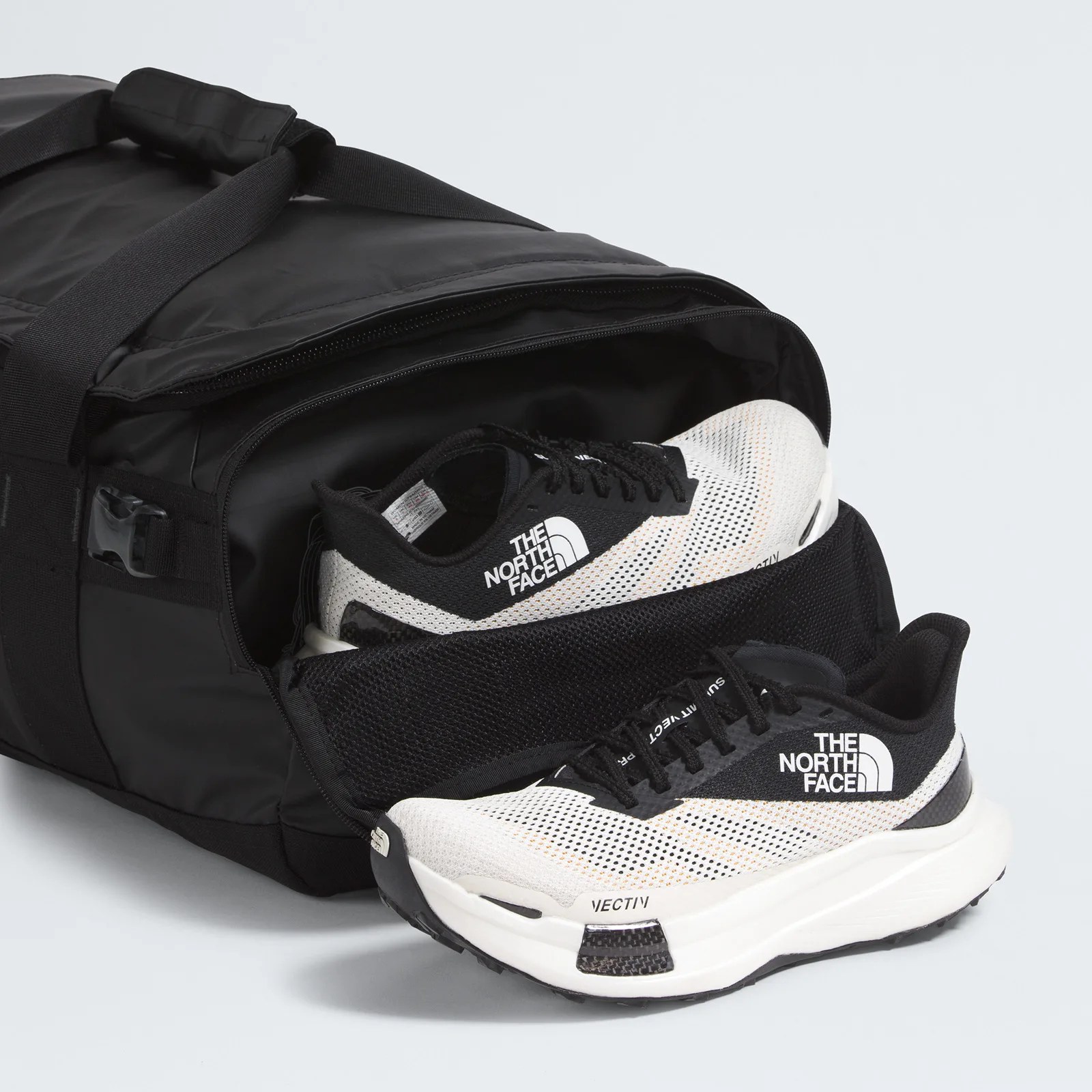 A pair of black and white The North Face VECTIV running shoes partially inside a black duffel bag with a zippered shoe compartment. The shoes feature a white mesh upper with black accents and the brand's logo prominently displayed on the side.