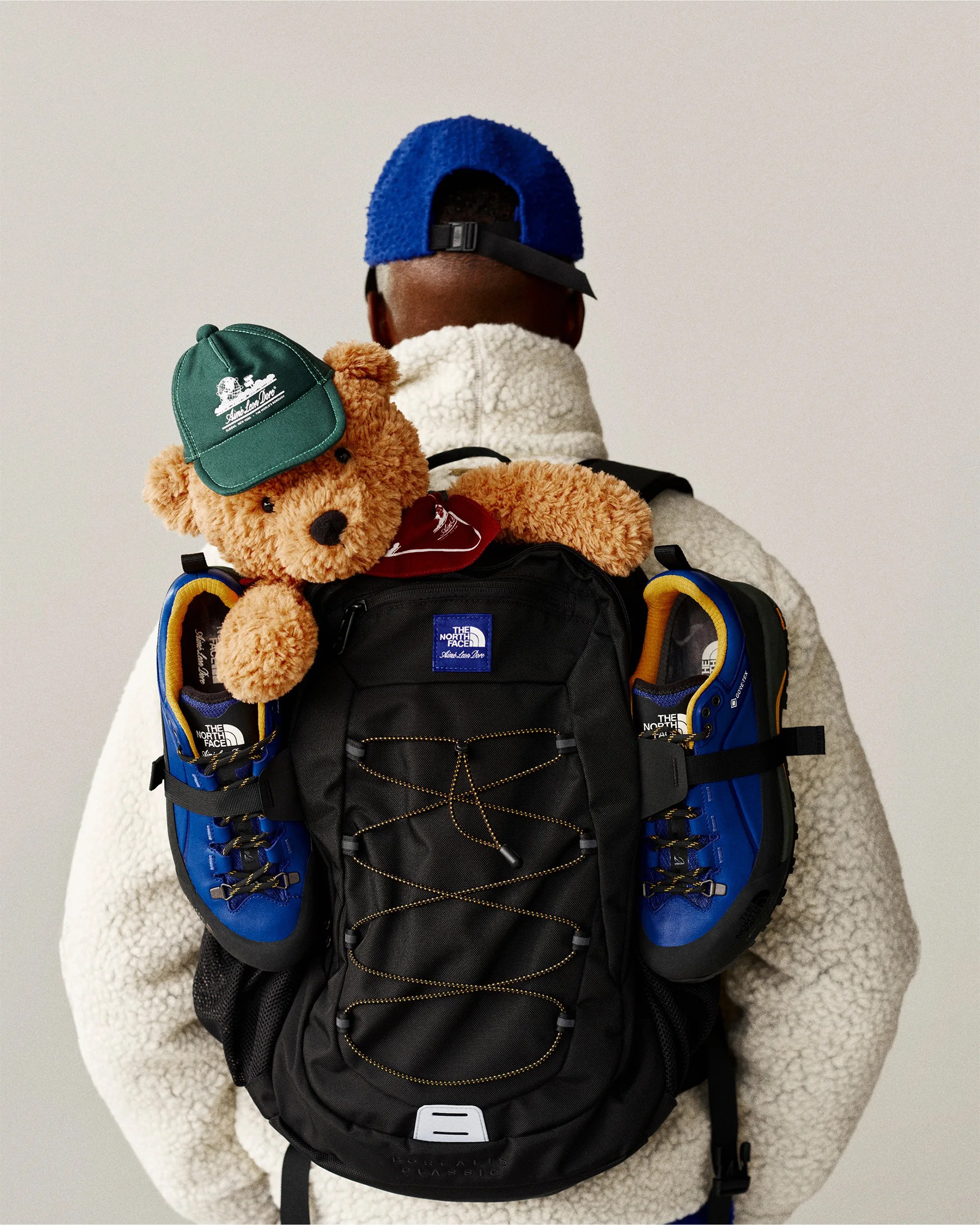 A person wearing a white fleece jacket and a blue cap is seen from behind, carrying a black The North Face backpack. Attached to the backpack are a pair of blue hiking shoes and a brown teddy bear wearing a green cap and a red bandana.