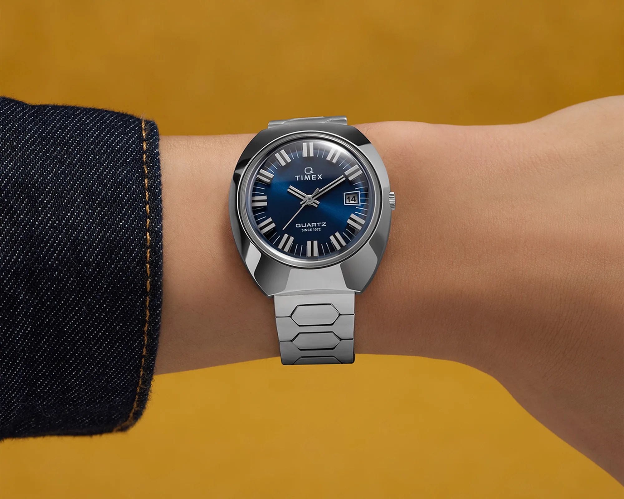 Silver Timex wristwatch with a blue dial, white hour markers, and a date display at the 3 o'clock position, worn on a wrist with a dark denim sleeve.