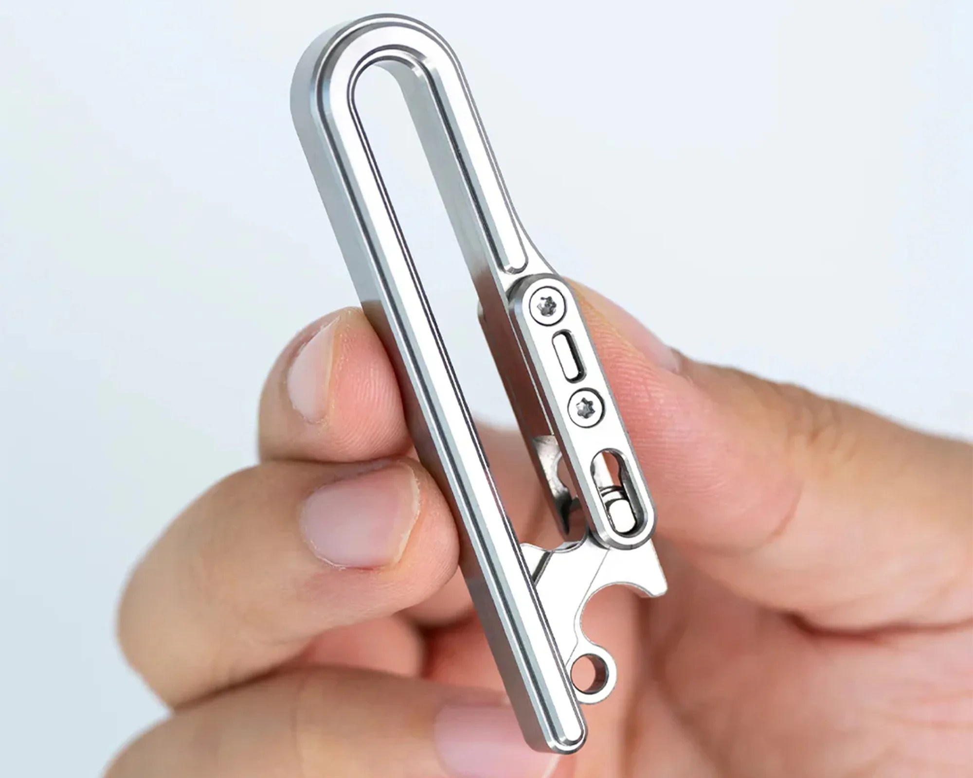 A close-up of a hand holding a sleek, silver metal clip with a minimalist design. The clip features two small screws and a sliding mechanism on one side, along with a small loop at the bottom, possibly for attaching keys or other items. The background is plain and light-colored.