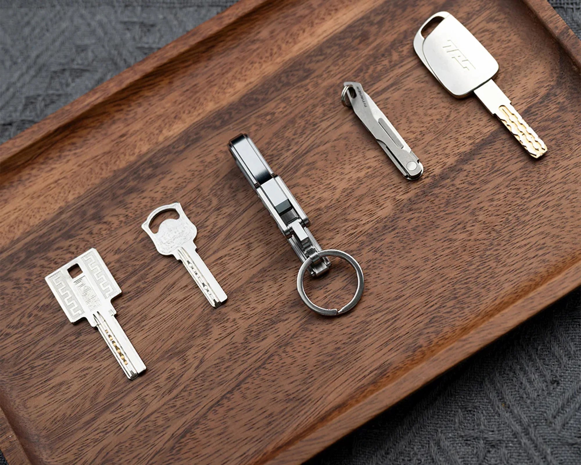 A wooden tray holding four silver keys and a small silver multi-tool with a keyring. The keys vary in shape and design, with one key having a rectangular head, another with a rounded head, and the others having unique designs. The multi-tool is compact and metallic, positioned in the center of the tray.
