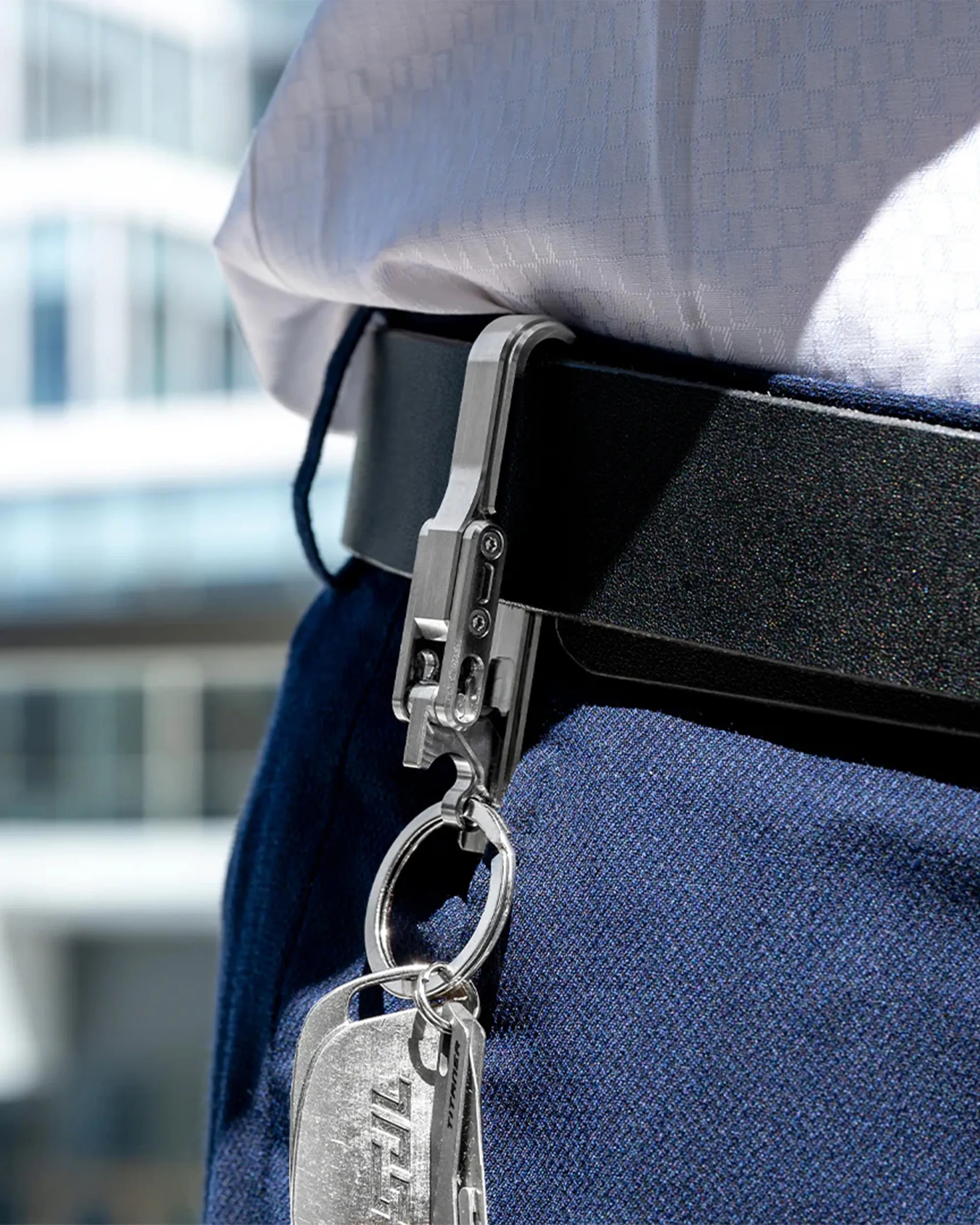 Metal key holder clip attached to a black belt, holding a keyring with two metal tags, worn with blue pants and a white shirt.