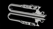 A close-up of a sleek, metallic folding tool or multi-tool with a polished finish, shown against a black background with a reflective surface beneath it. The tool features two parallel arms connected by screws and a small lever mechanism.