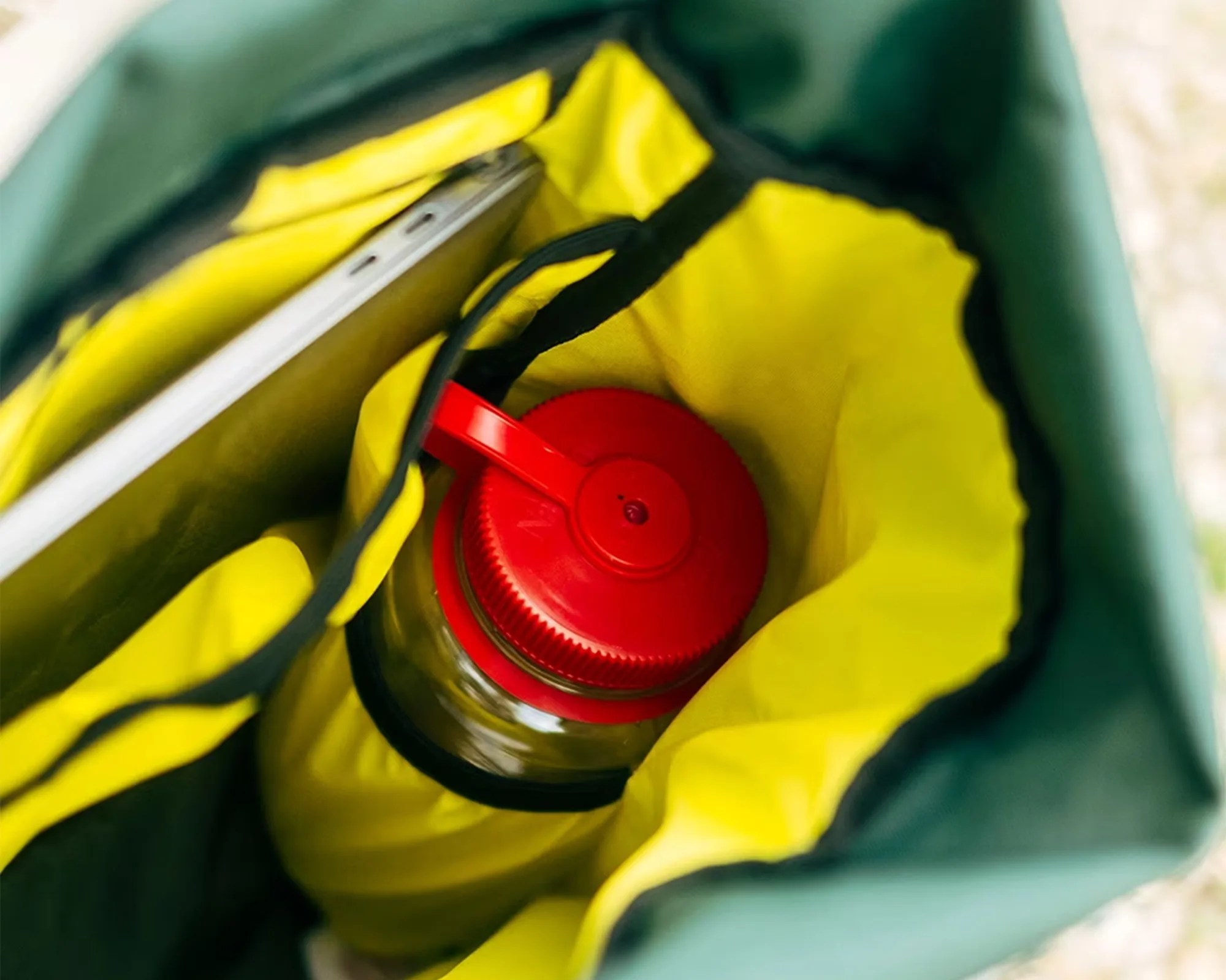Inside a green and yellow bag, there is a clear water bottle with a red cap and a silver laptop or tablet partially visible in a separate compartment.