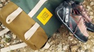 A green and brown Topo Designs backpack with white straps is lying on a rocky ground next to a pair of black and pink climbing shoes with red and white laces and the number "5.10" printed in white on the side.