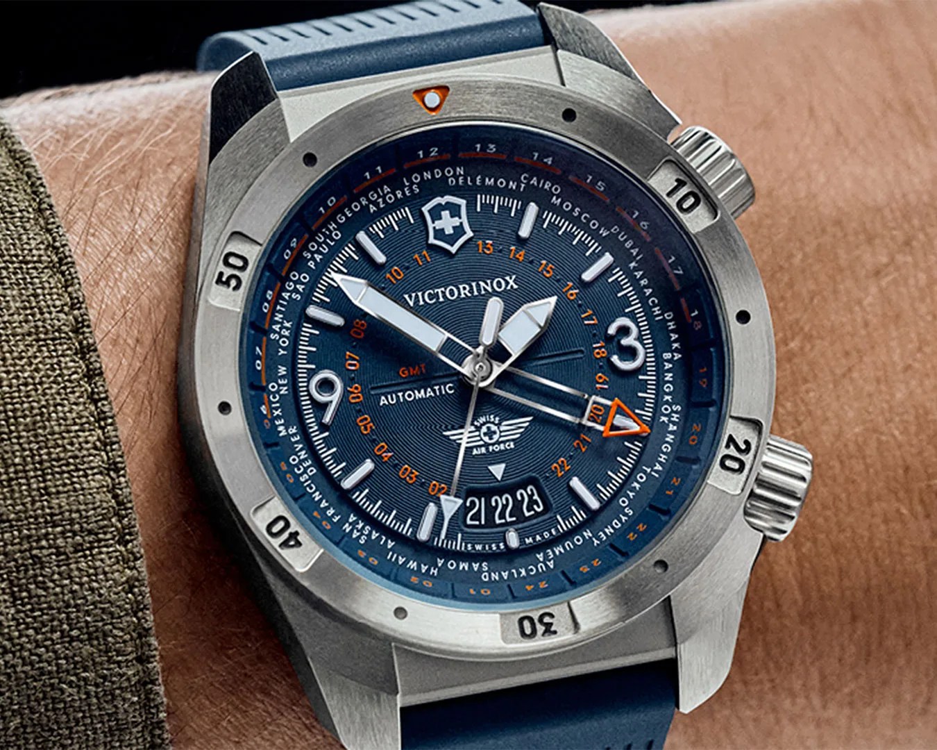 Victorinox wristwatch with a blue dial and silver stainless steel case, featuring large white hour markers and hands, an orange GMT hand, and a date display at the 6 o'clock position. The watch has a rotating bezel with minute markings and a blue rubber strap. The dial also includes city names for world time zones around the outer edge.