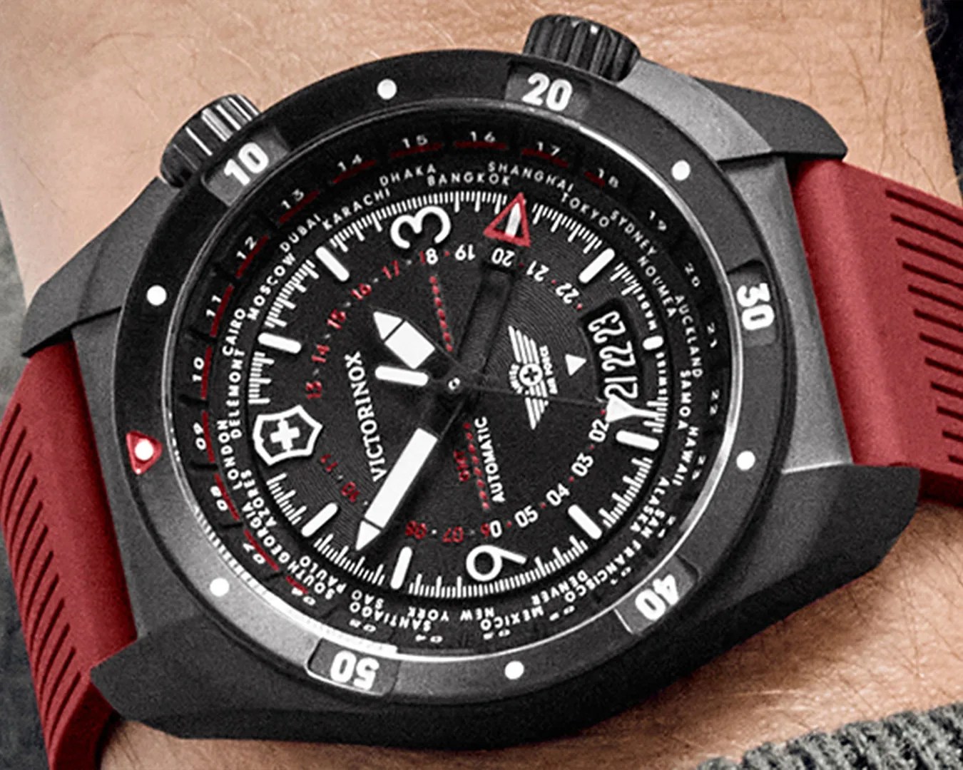 Black Victorinox automatic wristwatch with a red rubber strap, featuring a detailed dial with white and red markings, large white hour and minute hands, a red-tipped second hand, and city names around the outer edge for world time zones. The bezel is black with white numerals at 10, 20, 30, 40, and 50.