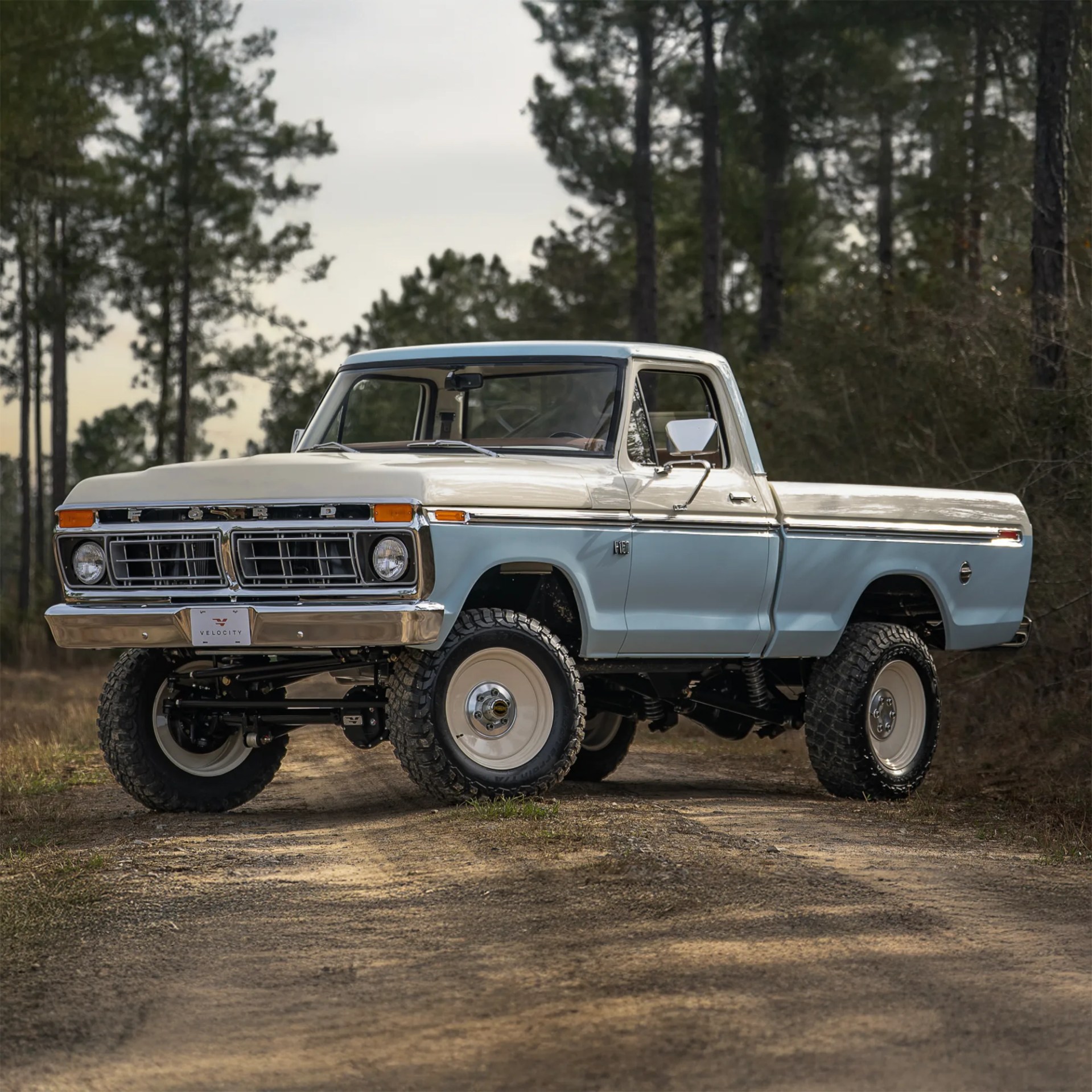 Light blue and white vintage Ford pickup truck with off-road tires parked on a dirt path surrounded by tall pine trees. The truck has a classic design with a chrome front grille and bumper.