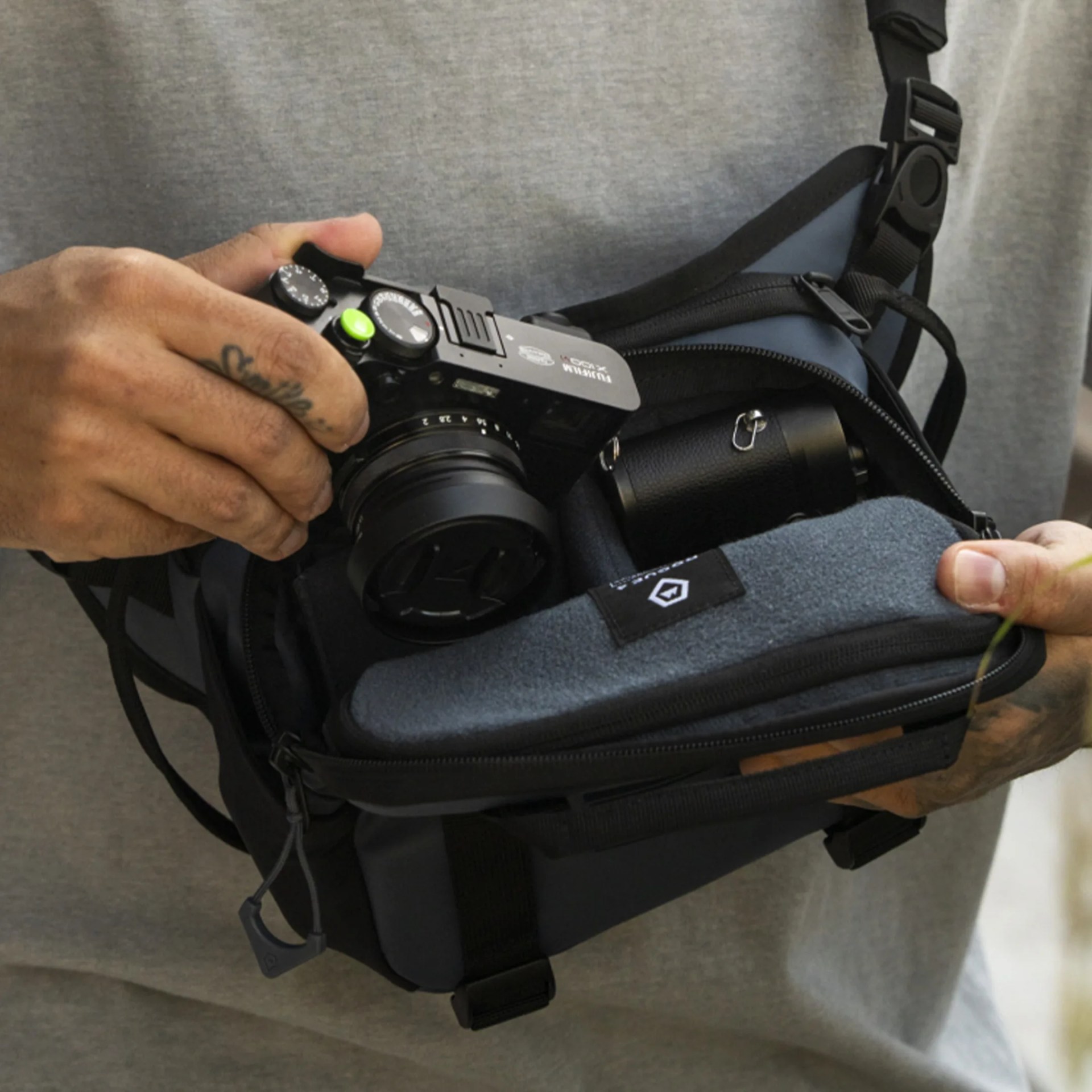 A person holding a black Fujifilm camera with a green shutter button, partially inside a black and gray camera bag. The bag is open, revealing a padded compartment and a black cylindrical object, likely a camera lens. The person is wearing a gray shirt and has a tattoo on their left hand.