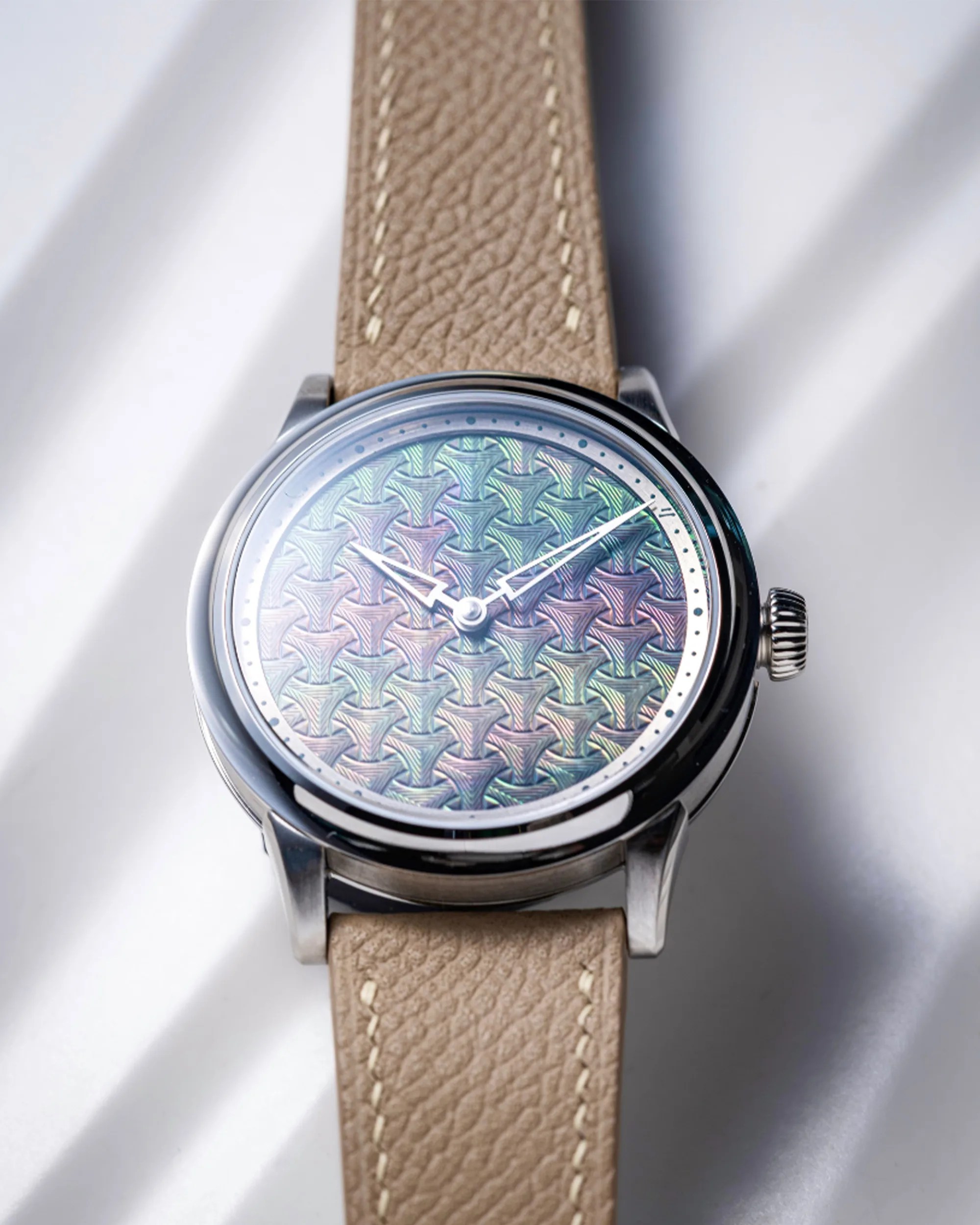 A wristwatch with a polished silver case and a beige leather strap featuring white stitching. The watch face has a unique iridescent pattern with geometric shapes in shades of green, purple, and pink. The watch hands are silver with a distinctive design, and there are small dots marking the hours around the edge of the dial.