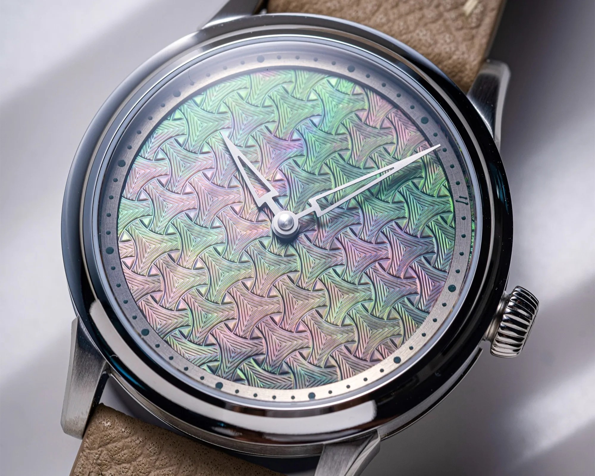 Close-up of a wristwatch with a polished silver case and a beige leather strap. The watch face features an intricate, multicolored geometric pattern with interlocking shapes in pastel shades of green, pink, and purple. The watch hands are silver with a skeleton design, and there are small black dots marking the minutes around the edge of the dial.