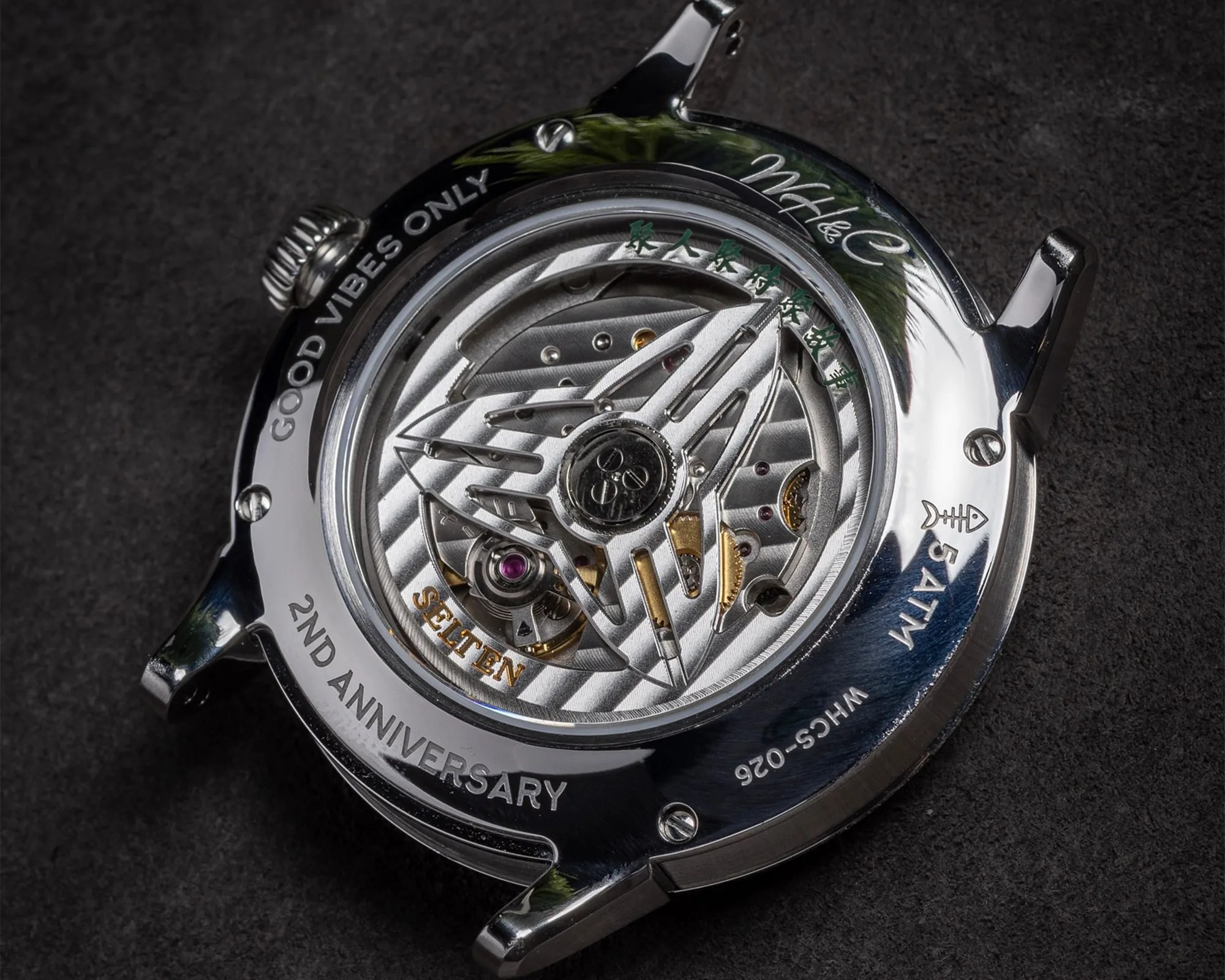 Back view of a mechanical watch case showcasing the intricate movement inside. The rotor features a star-shaped design with the word "SELTEN" in gold lettering. The polished metal case has engravings including "GOOD VIBES ONLY," "2ND ANNIVERSARY," "5 ATM," and "WHCS-026." There are also some green Chinese characters and a signature near the top right. The watch is placed on a dark textured surface.