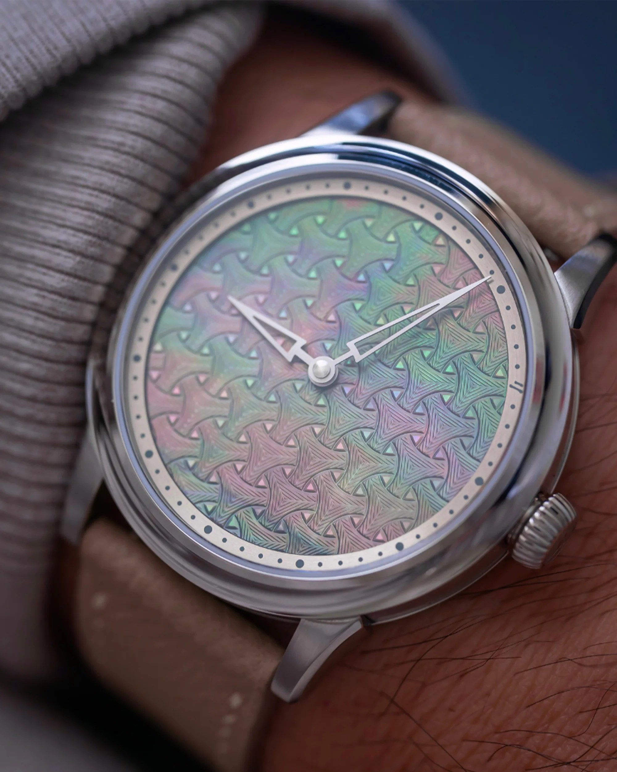 Close-up of a wristwatch with a silver case and a beige leather strap. The watch face features an intricate, multicolored geometric pattern with interlocking shapes in pastel shades of green, pink, and purple. The watch hands are silver with a skeleton design, and small black dots mark the hours around the edge of the dial.