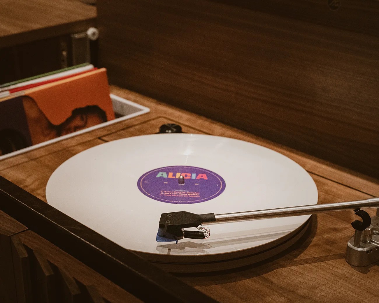 A white vinyl record labeled "ALICIA" with a purple center is playing on a wooden turntable. The tonearm and needle are positioned on the record, and there are several other vinyl records stored in a compartment next to the turntable. The setup has a warm, vintage aesthetic with wood surfaces.