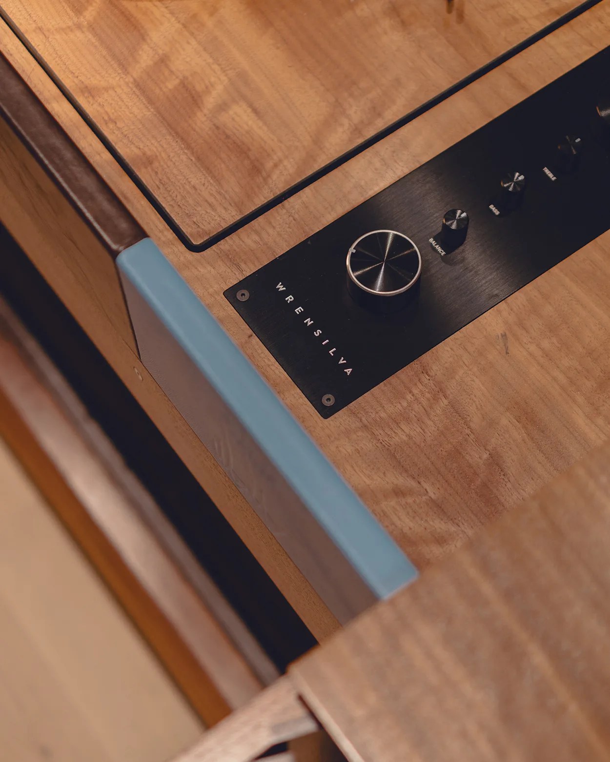 Close-up of a wooden audio control panel featuring a large silver knob and smaller black knobs labeled "Balance," "Bass," and "Treble." The panel has the brand name "Wrensilva" printed on it. The wood has a smooth, natural finish, and there is a blue and brown edge detail visible on the side.